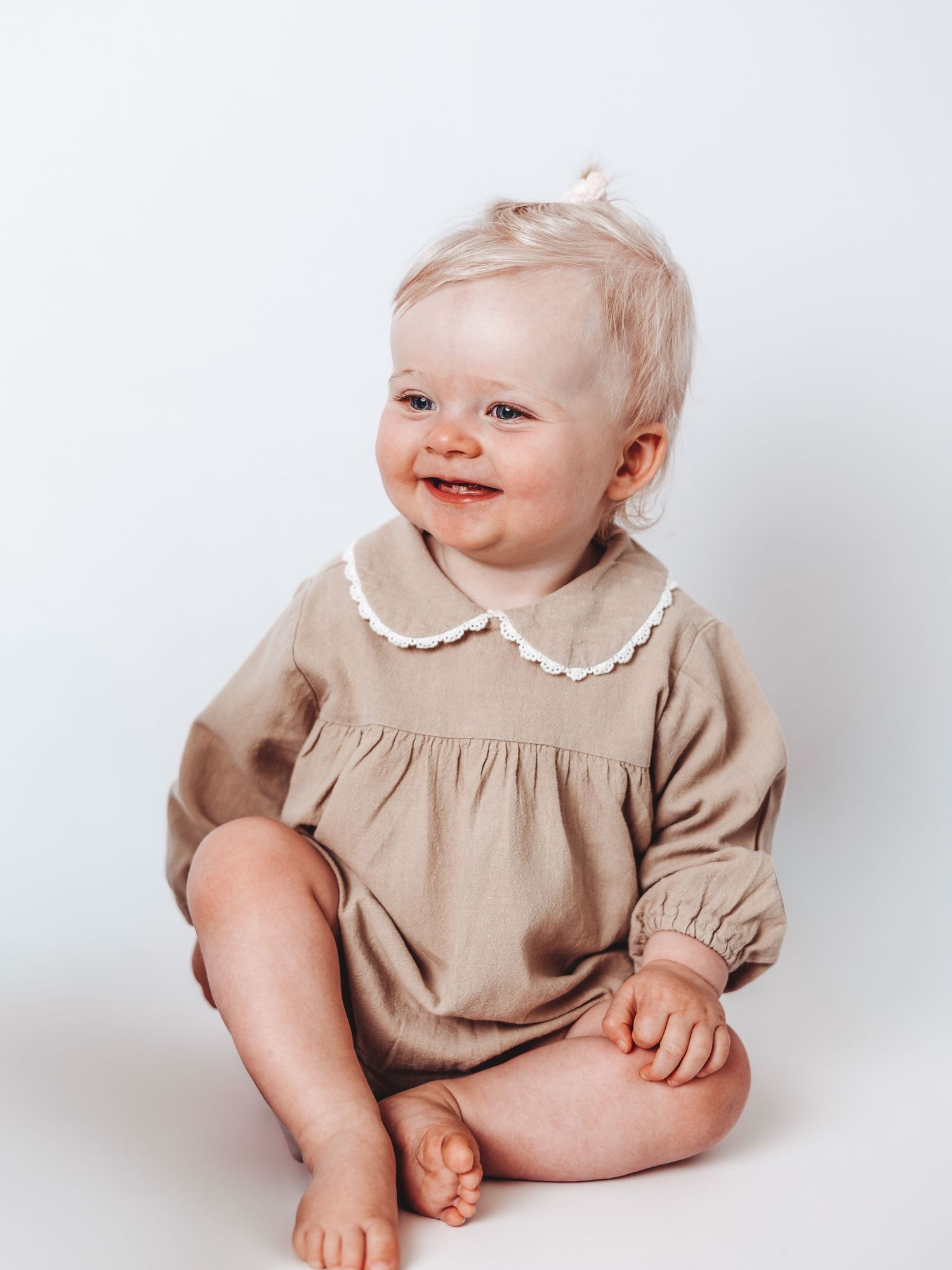 A smiling baby wearing a beige dress with peter pan collar sits on a white background.
