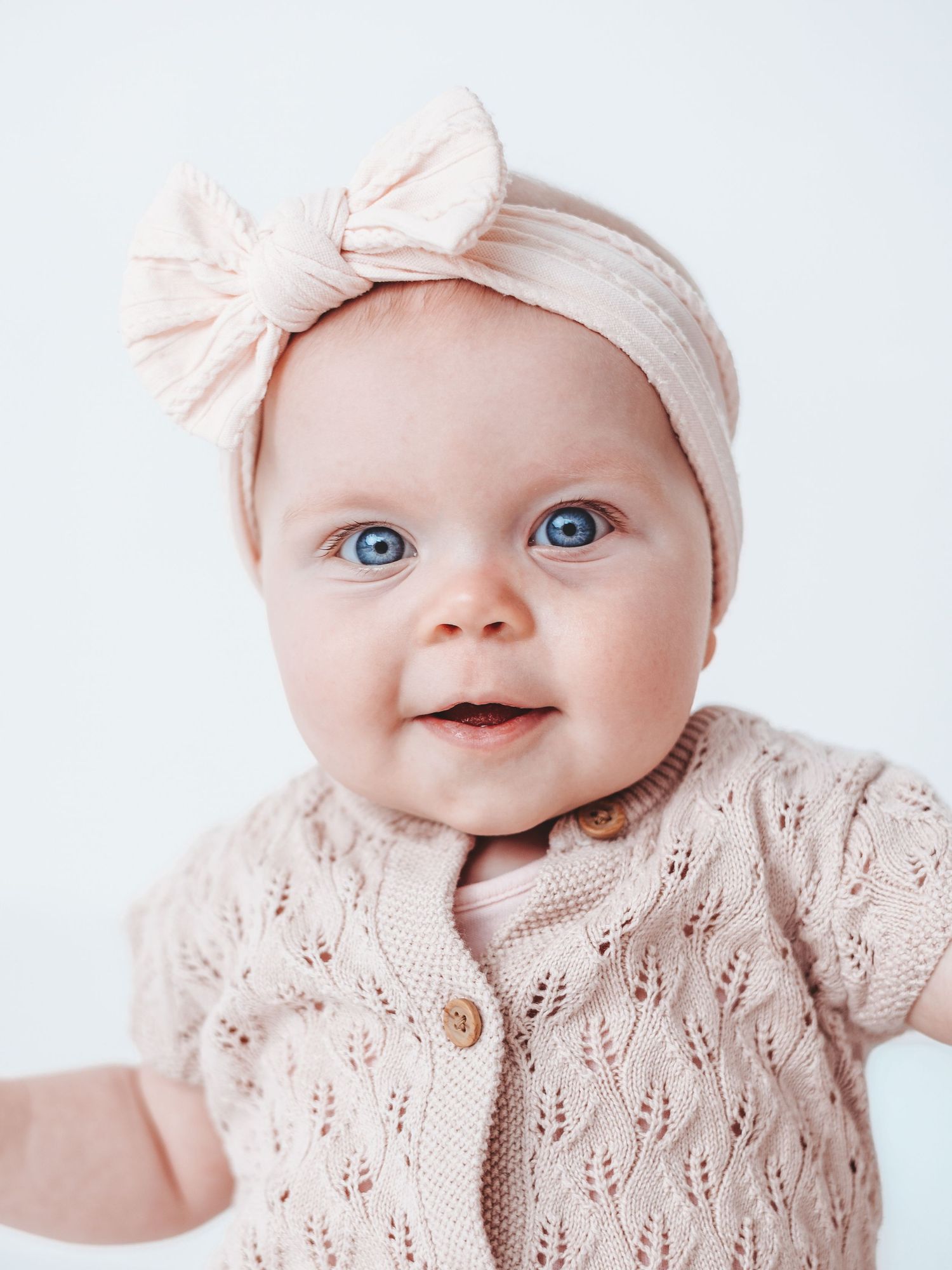 Baby wearing pink knit cardigan and matching bow headband smiles while posing against white background.
