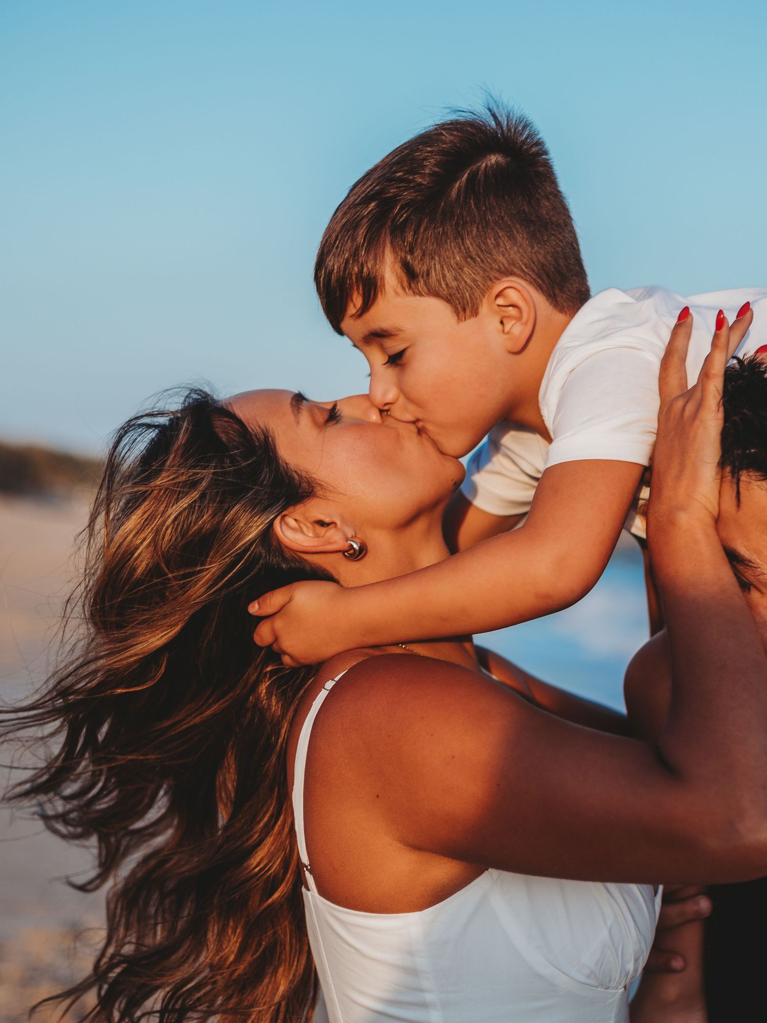A mother lovingly kisses her young son on the beach at sunset, holding him up in her arms.