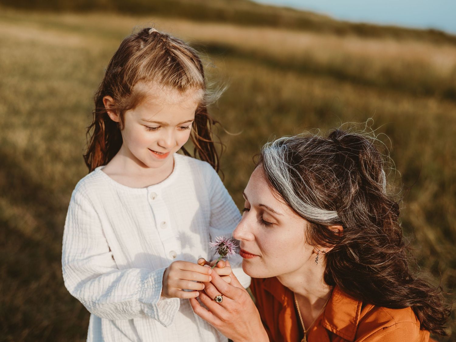 A mother and young daughter share a tender moment smelling wildflowers together in a sunlit golden field.