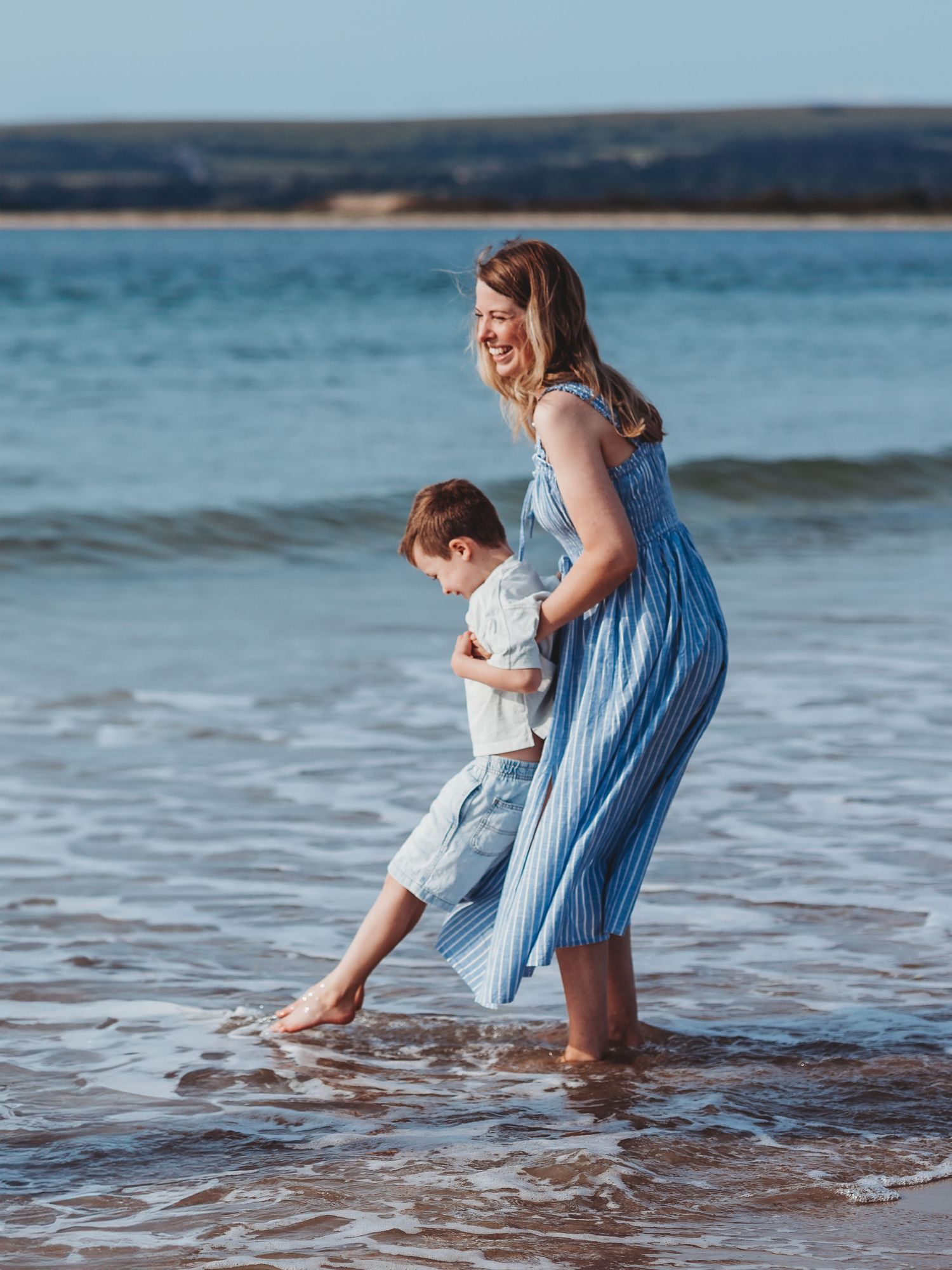 A smiling woman in a blue striped dress plays with a young boy in shallow ocean waves on a sunny beach.