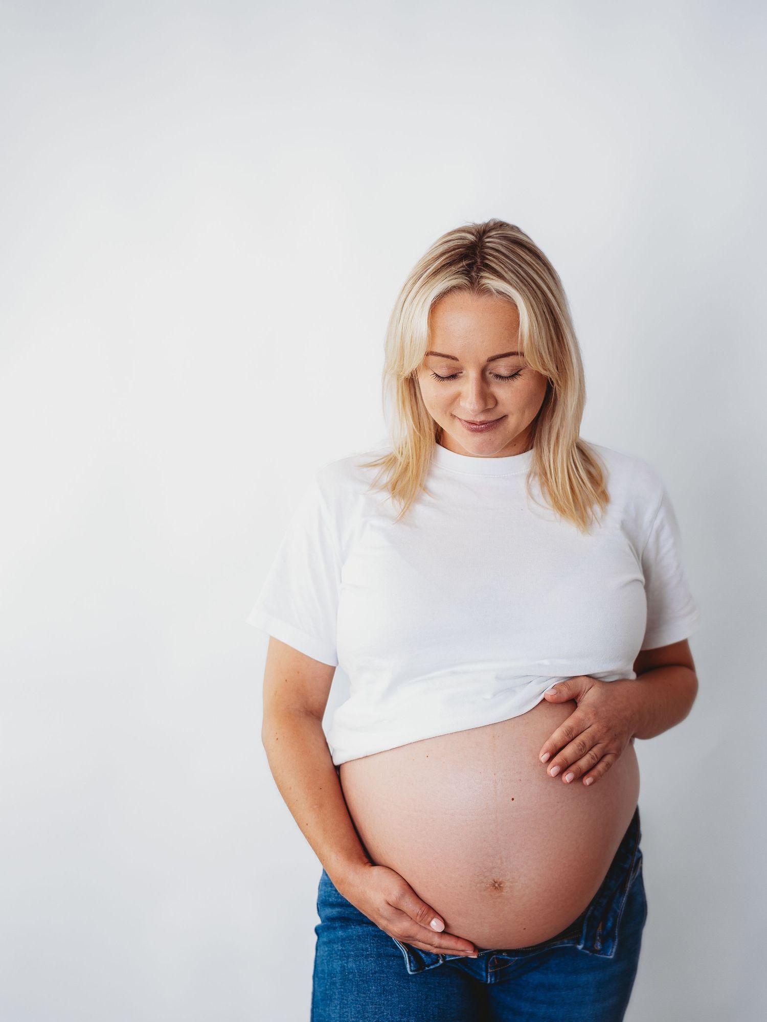 A pregnant woman in a white t-shirt and jeans poses against a light background showing her growing belly.