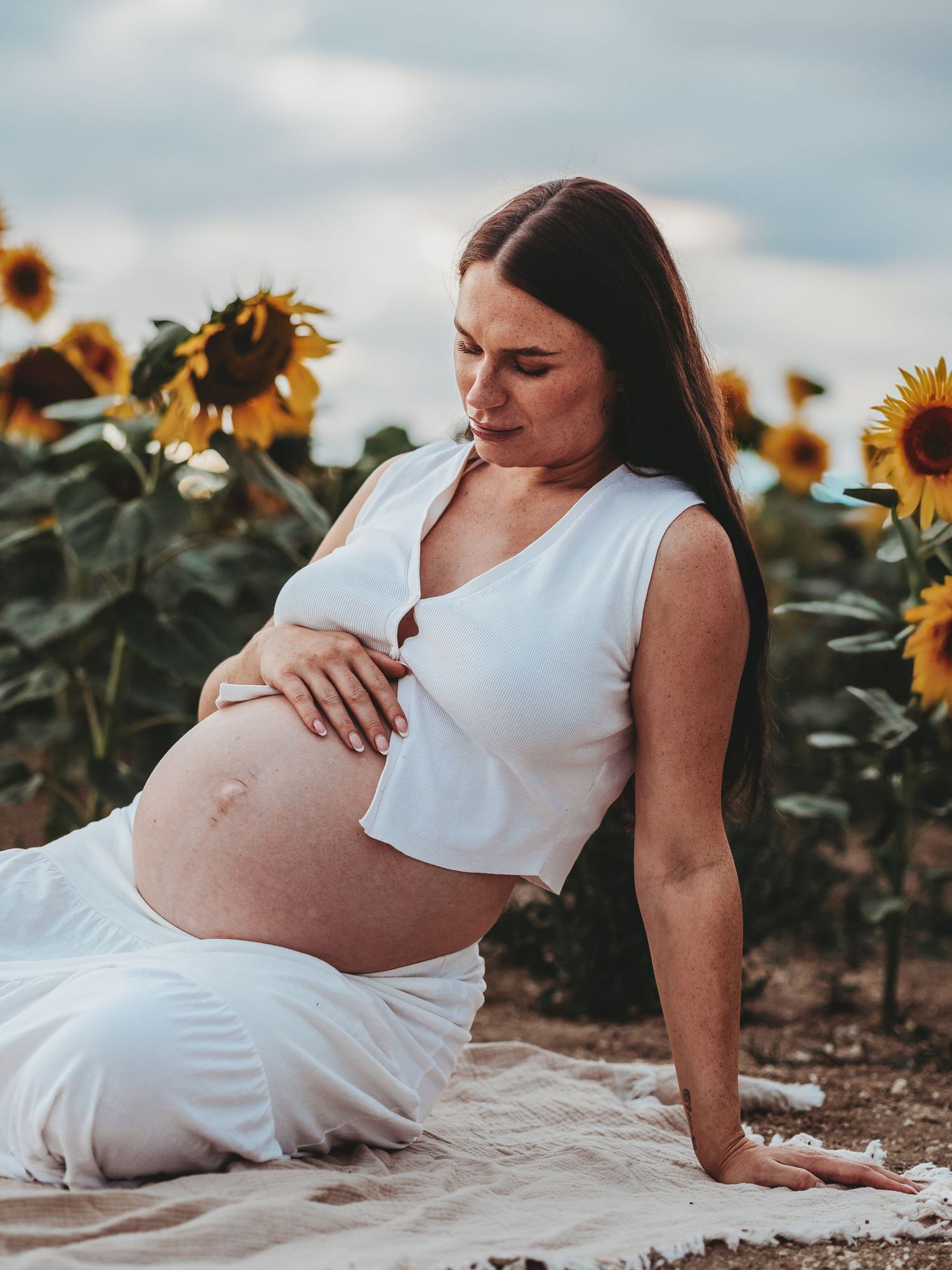 Maternity photoshoot in a sunflower field shows a woman in white dress reclining on ground during golden hour.
