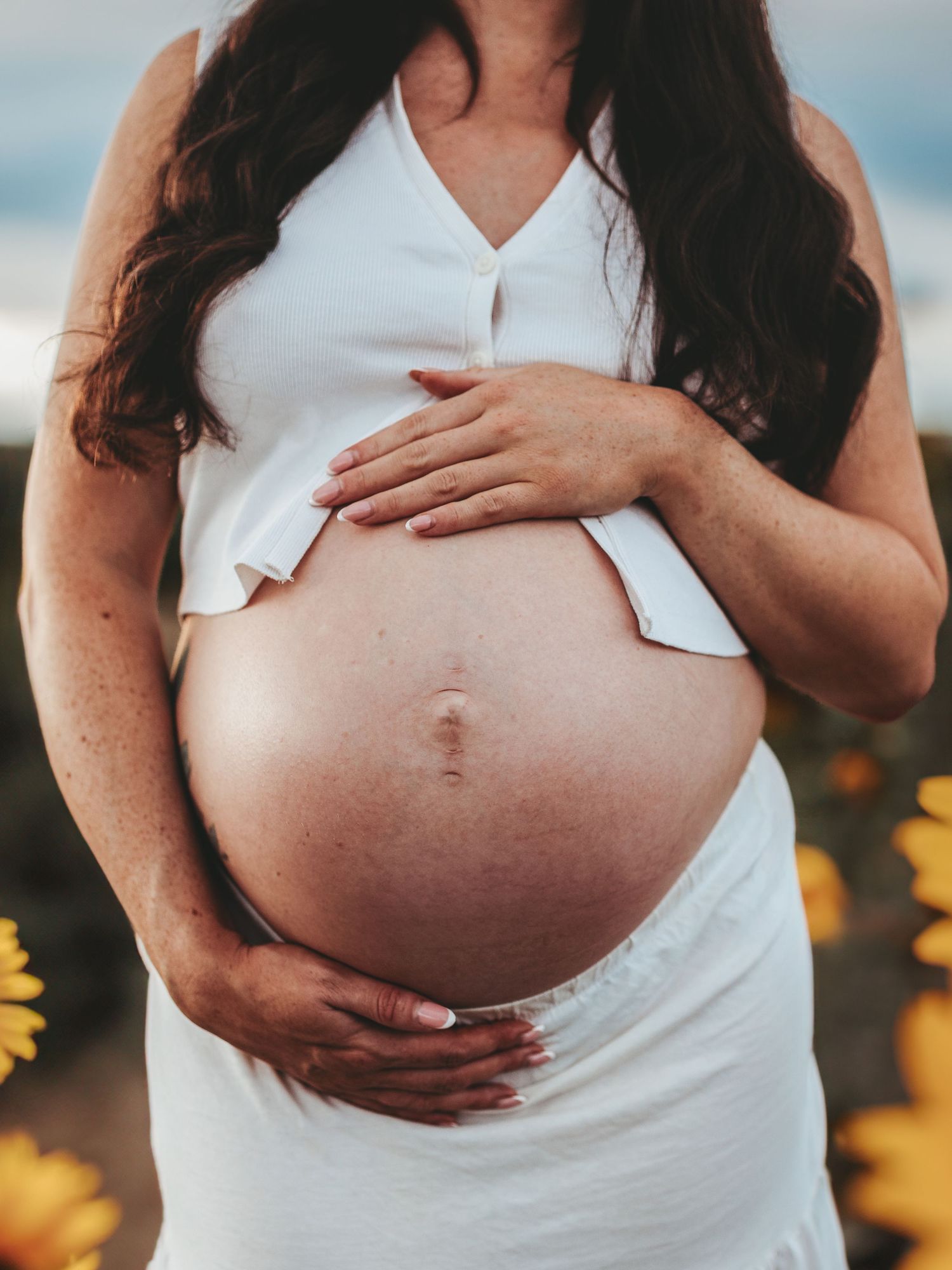 A pregnant woman in a white dress cradles her belly while standing in front of blooming sunflowers.