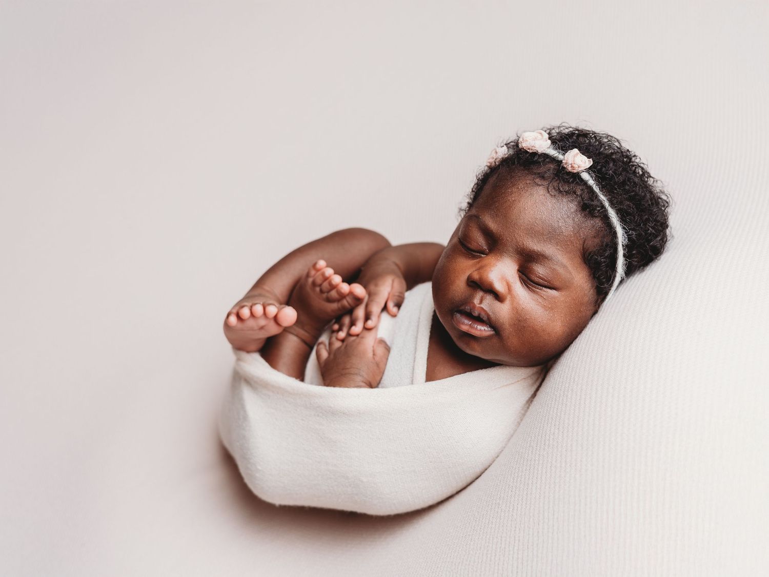 A sleeping newborn baby wrapped in white fabric lies peacefully against a light colored background.