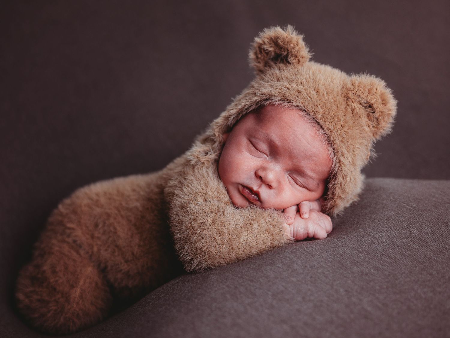 A sleeping newborn wearing a soft brown teddy bear outfit rests peacefully on a dark gray cushion.