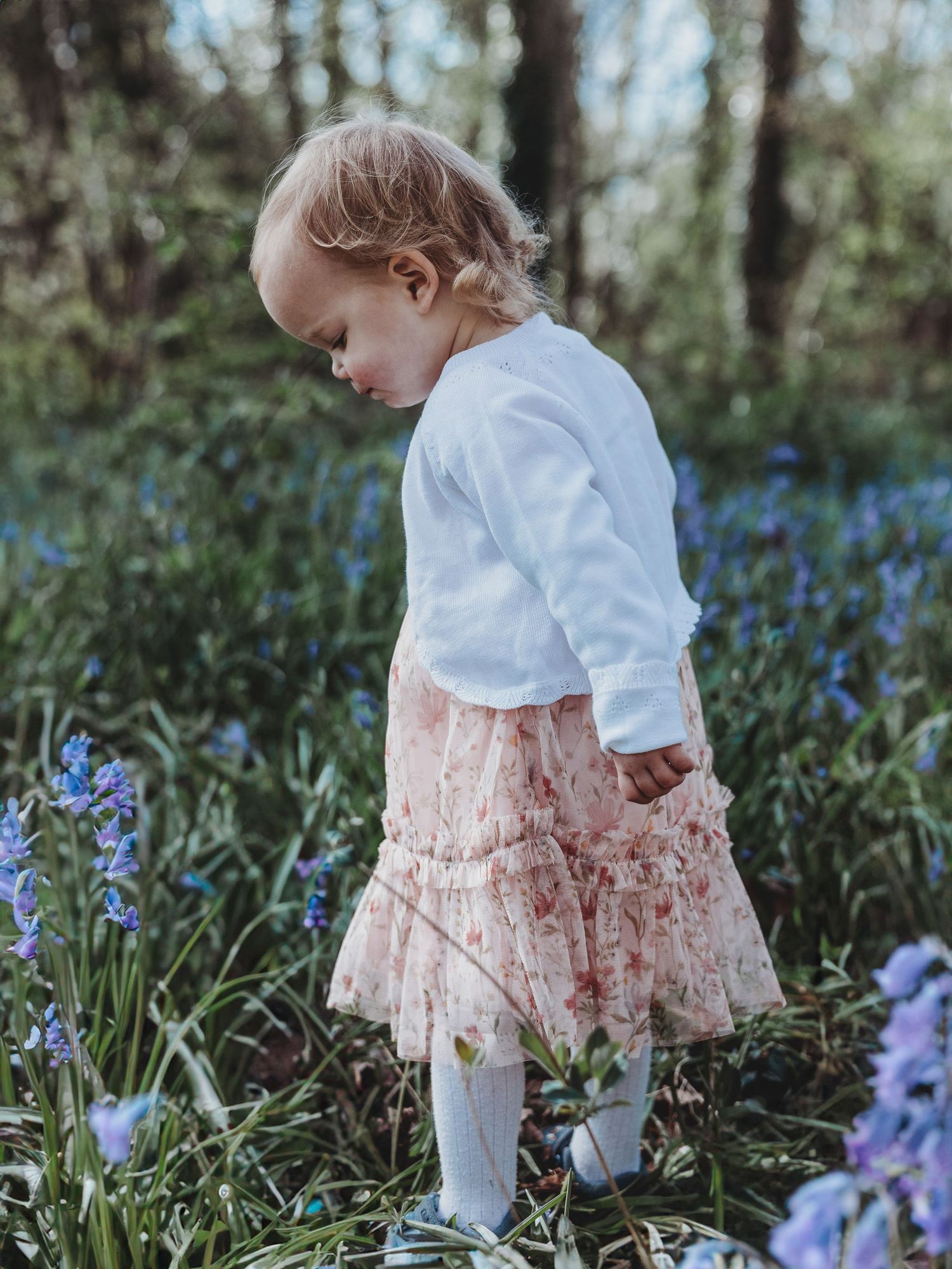 Toddler girl in white top and pink floral skirt exploring bluebell flowers in a woodland garden.