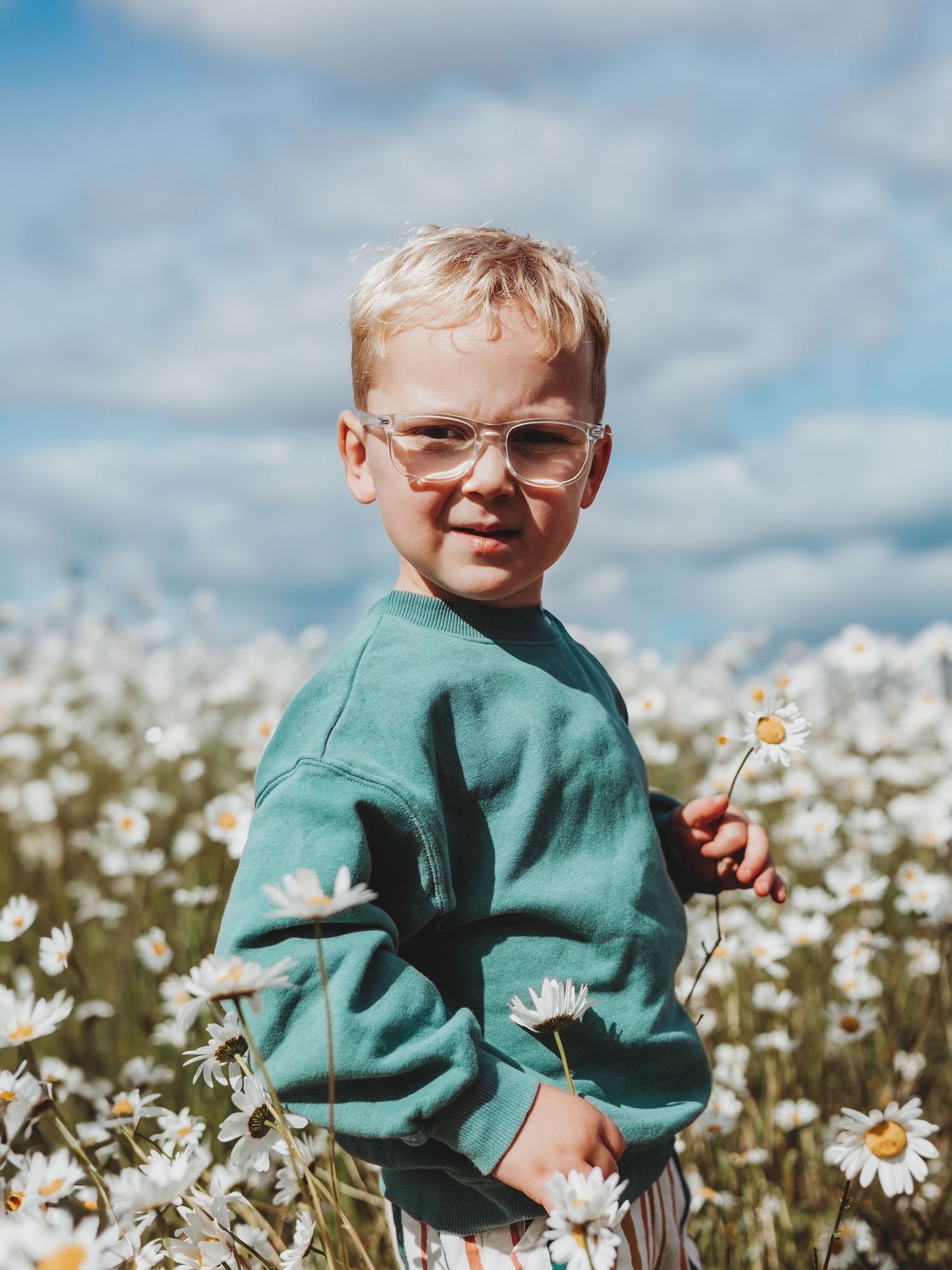 A young blonde boy wearing glasses and a teal sweatshirt stands in a field of white daisies under a blue sky.