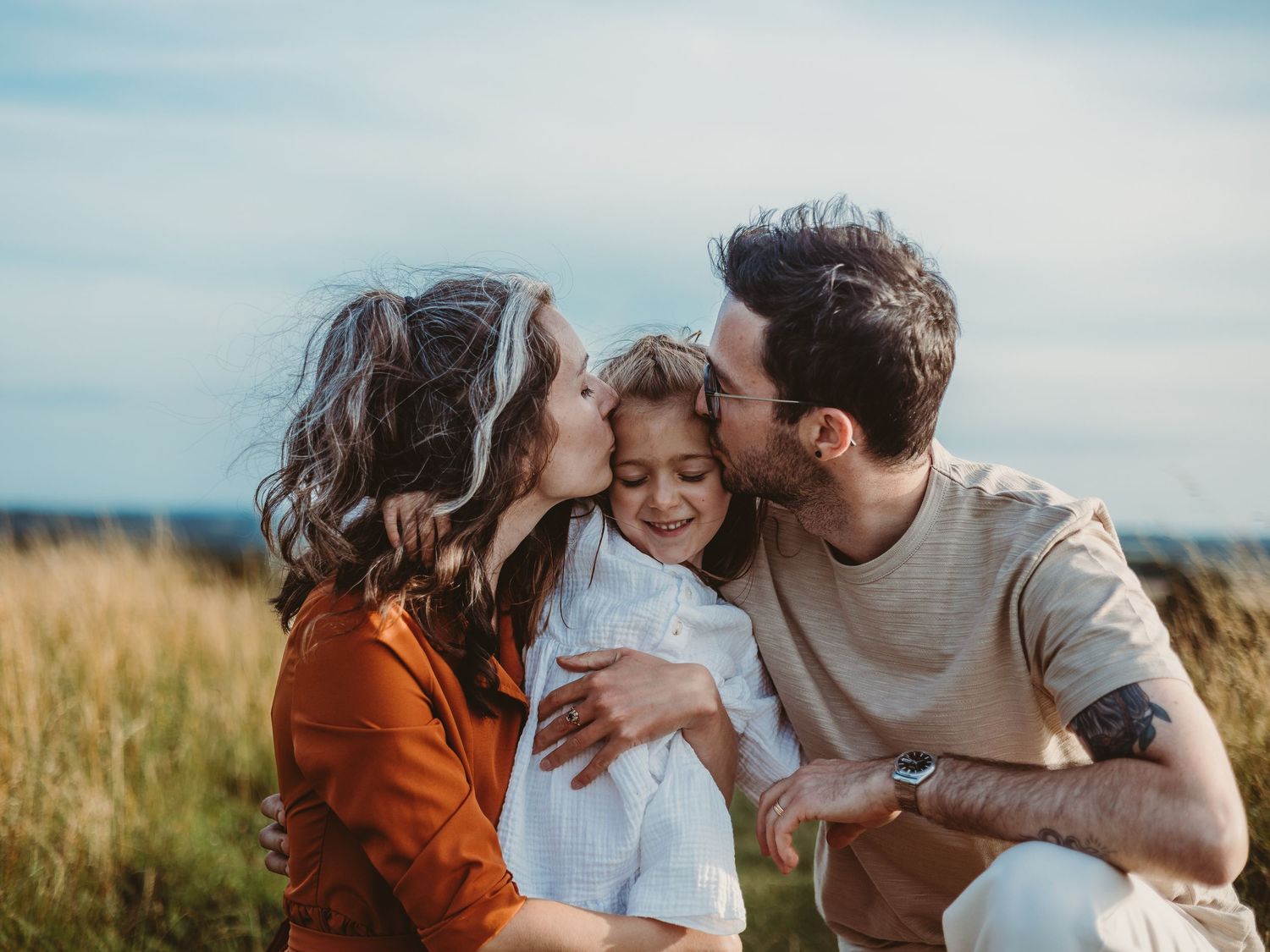 Parents kissing their smiling young daughter on the cheeks in a sunny golden field outdoors.