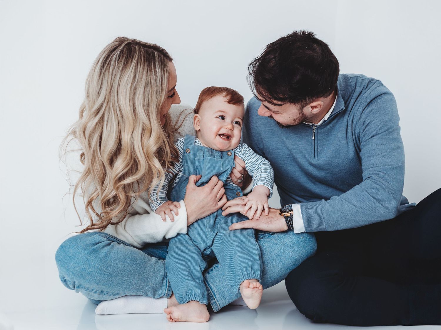 Happy family with blonde mother, dark-haired father, and smiling baby in blue outfits sitting on white background.
