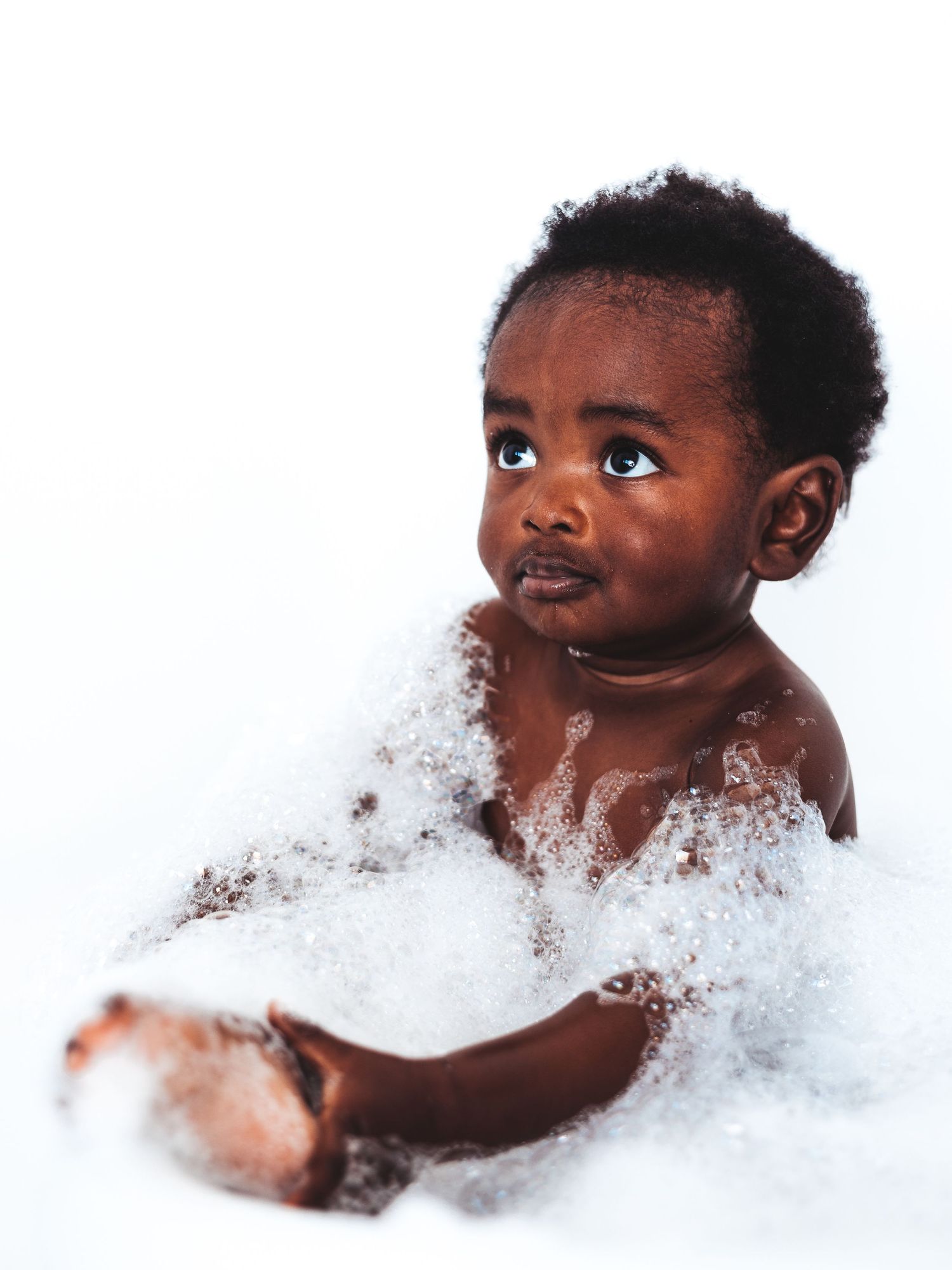 Adorable Black baby sitting in a bubble bath, looking up curiously with wide eyes against a white background.
