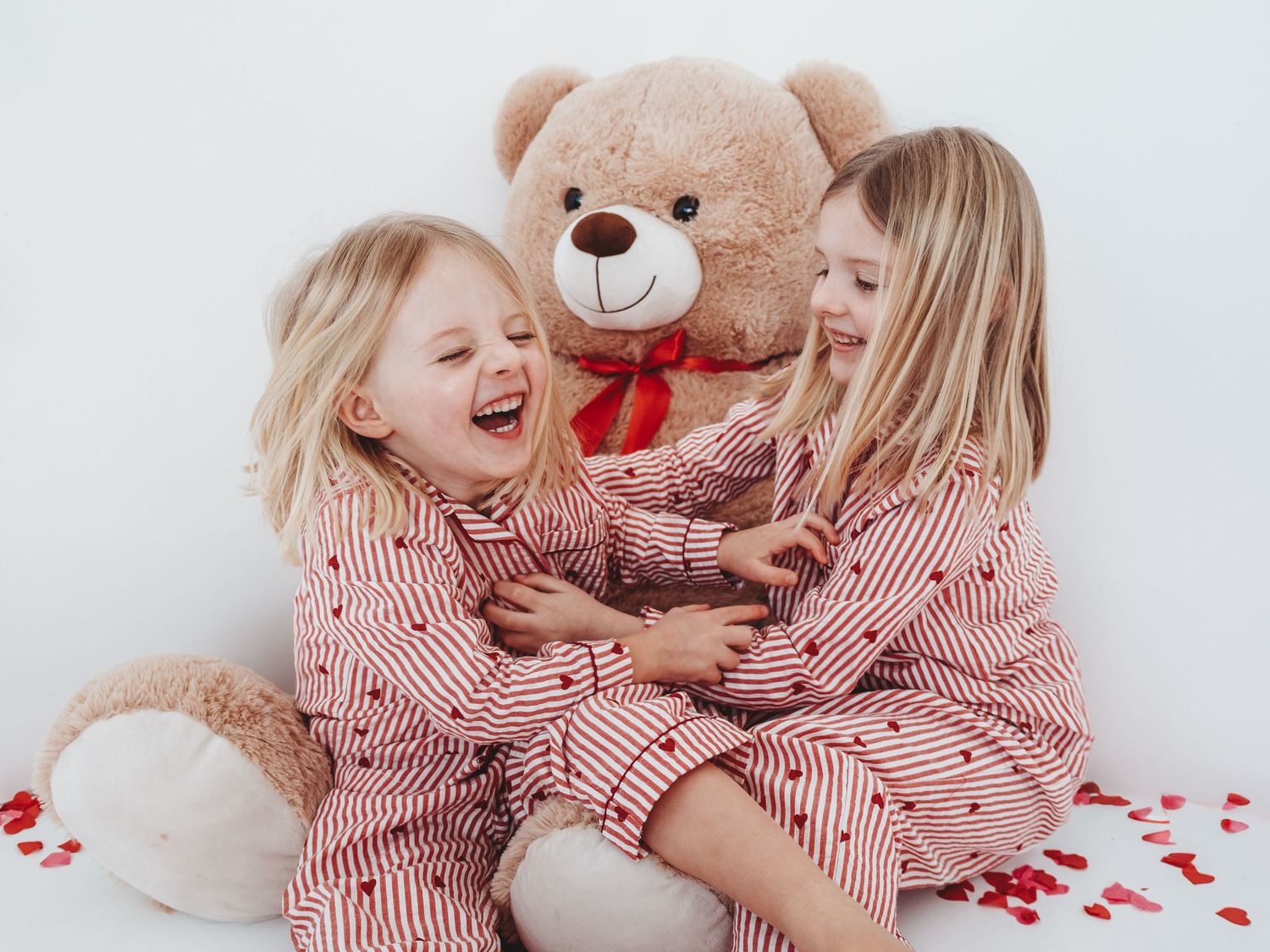 Children in matching red striped pajamas laugh and play with a large teddy bear on white bedding.
