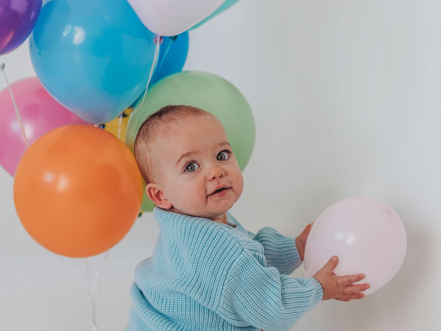A baby in a blue striped onesie plays with colorful balloons against a white background.
