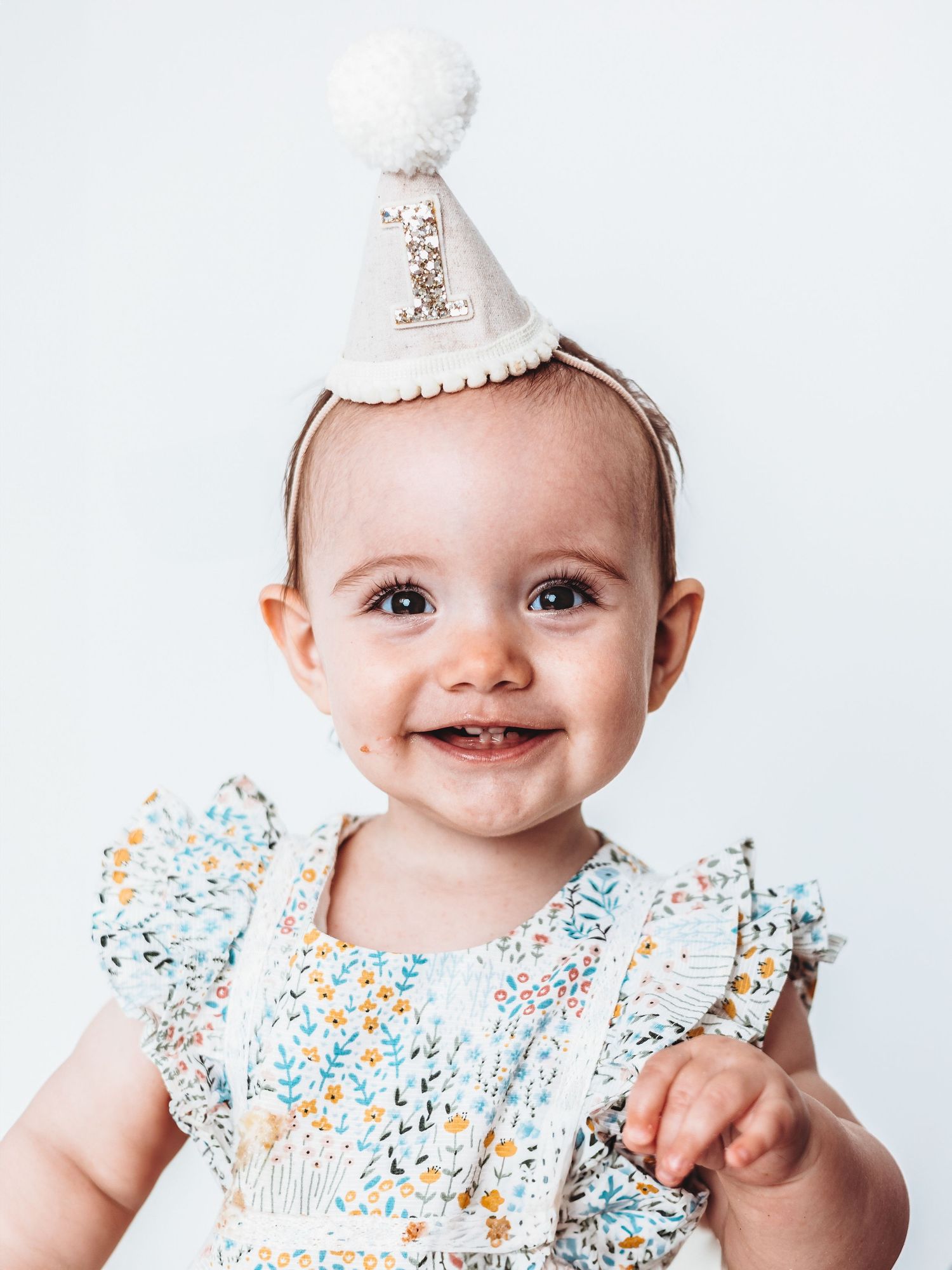 Smiling baby girl in floral romper wearing a white number 1 birthday party hat celebrating her first birthday.