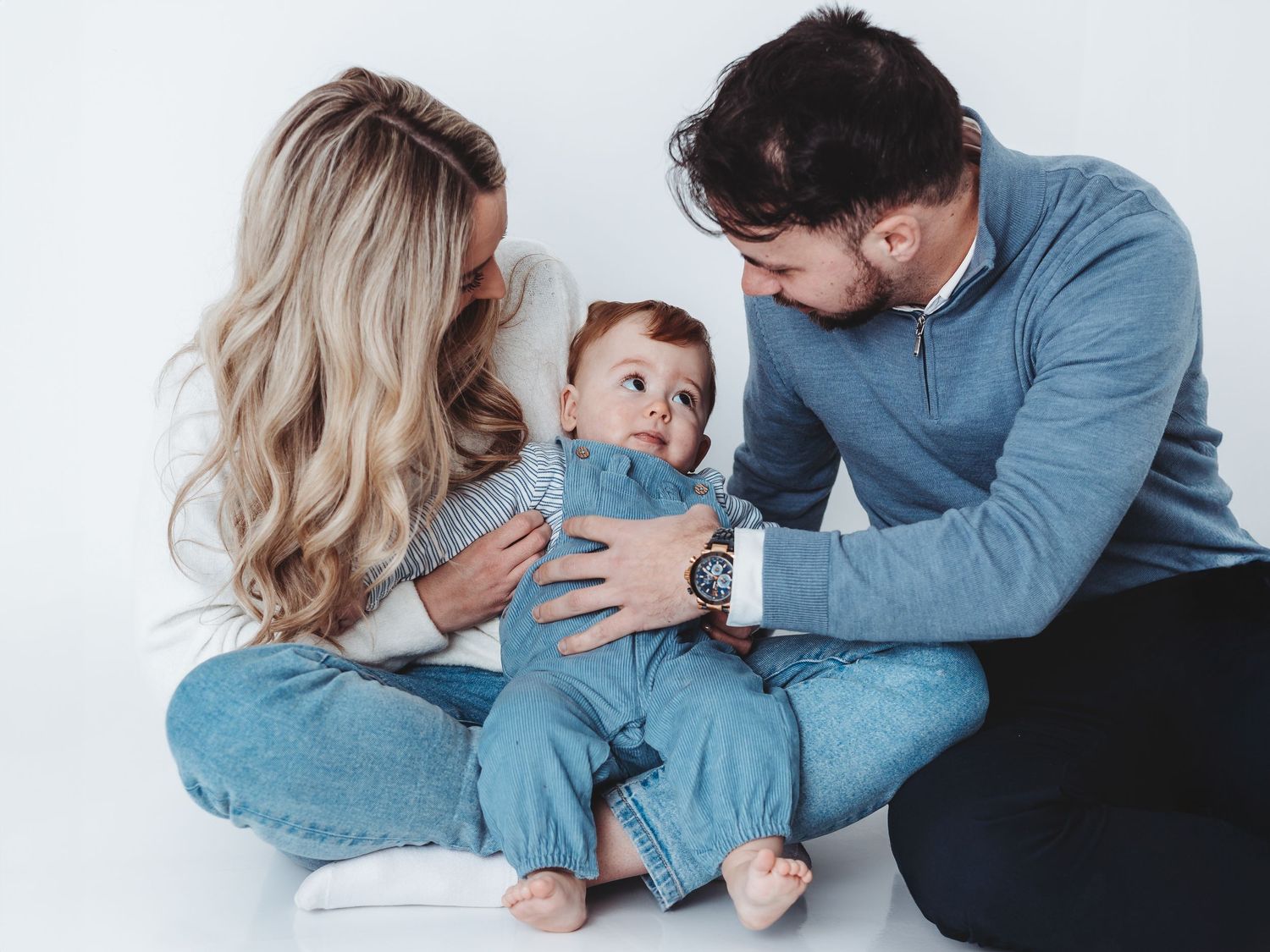 Young family with blonde mother and dark-haired father sitting on floor smiling at their baby in blue outfit.