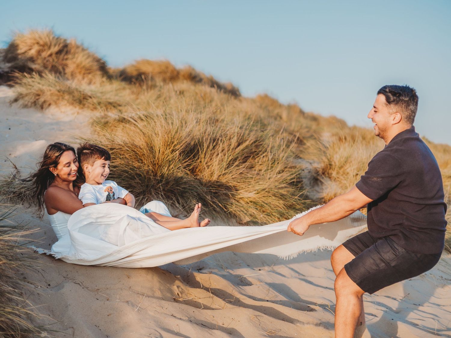Family enjoying a playful moment on sandy beach dunes, with parents and baby wrapped in white blanket at golden hour.