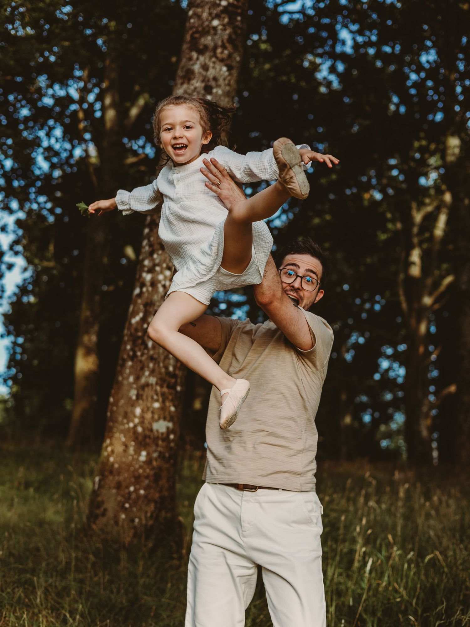 A joyful man lifting a laughing little girl high in the air outdoors in a sunlit forest setting.