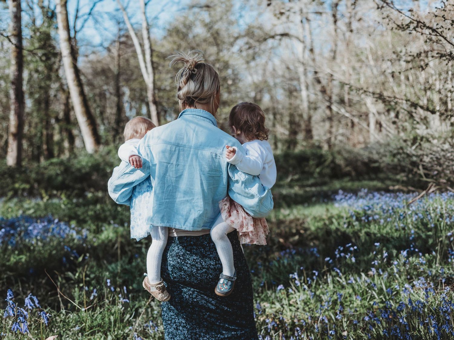 A mother carrying two young children through a bluebell woodland in spring, viewed from behind.
