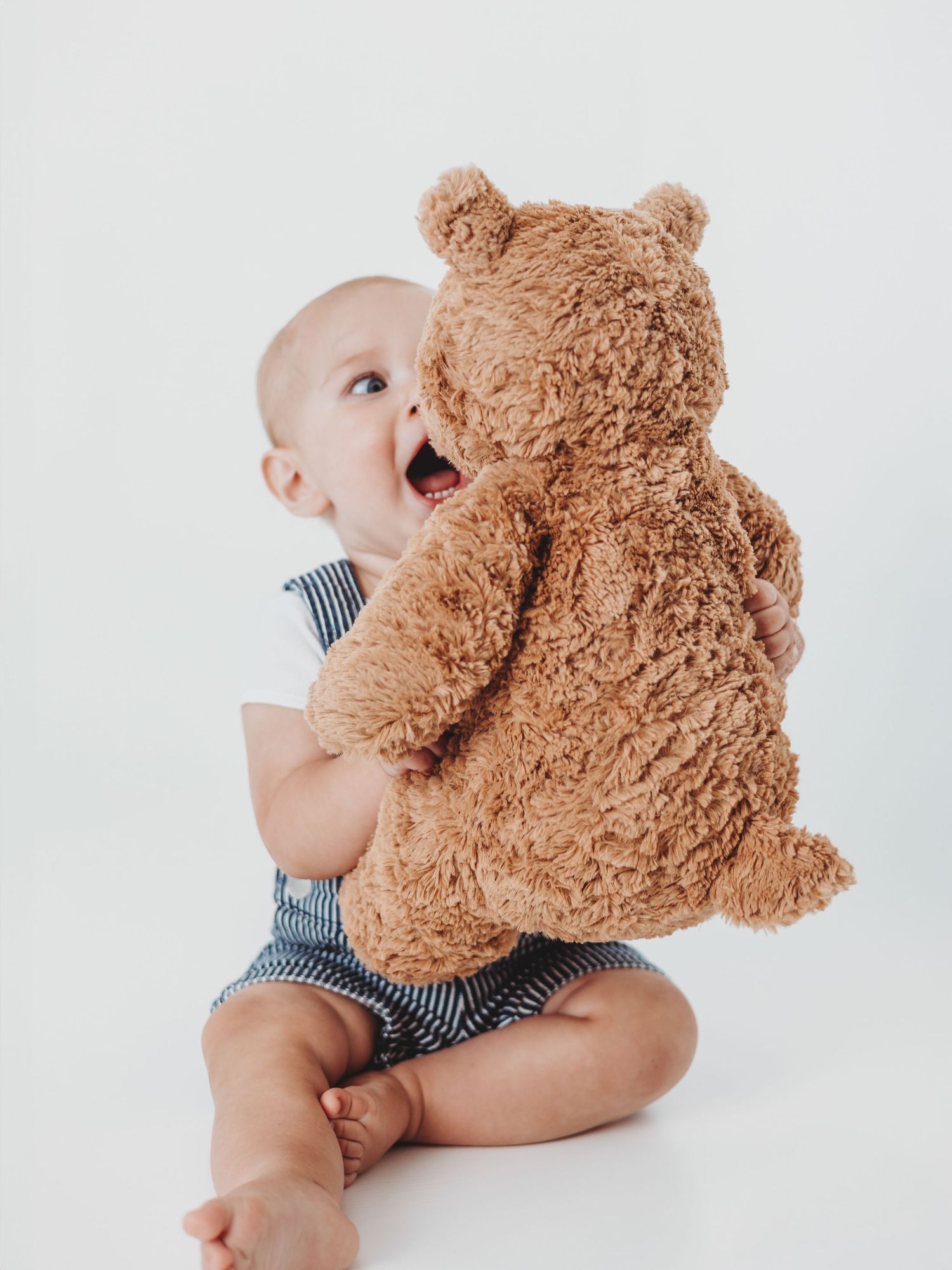 Baby in striped romper plays with large brown teddy bear against white background.