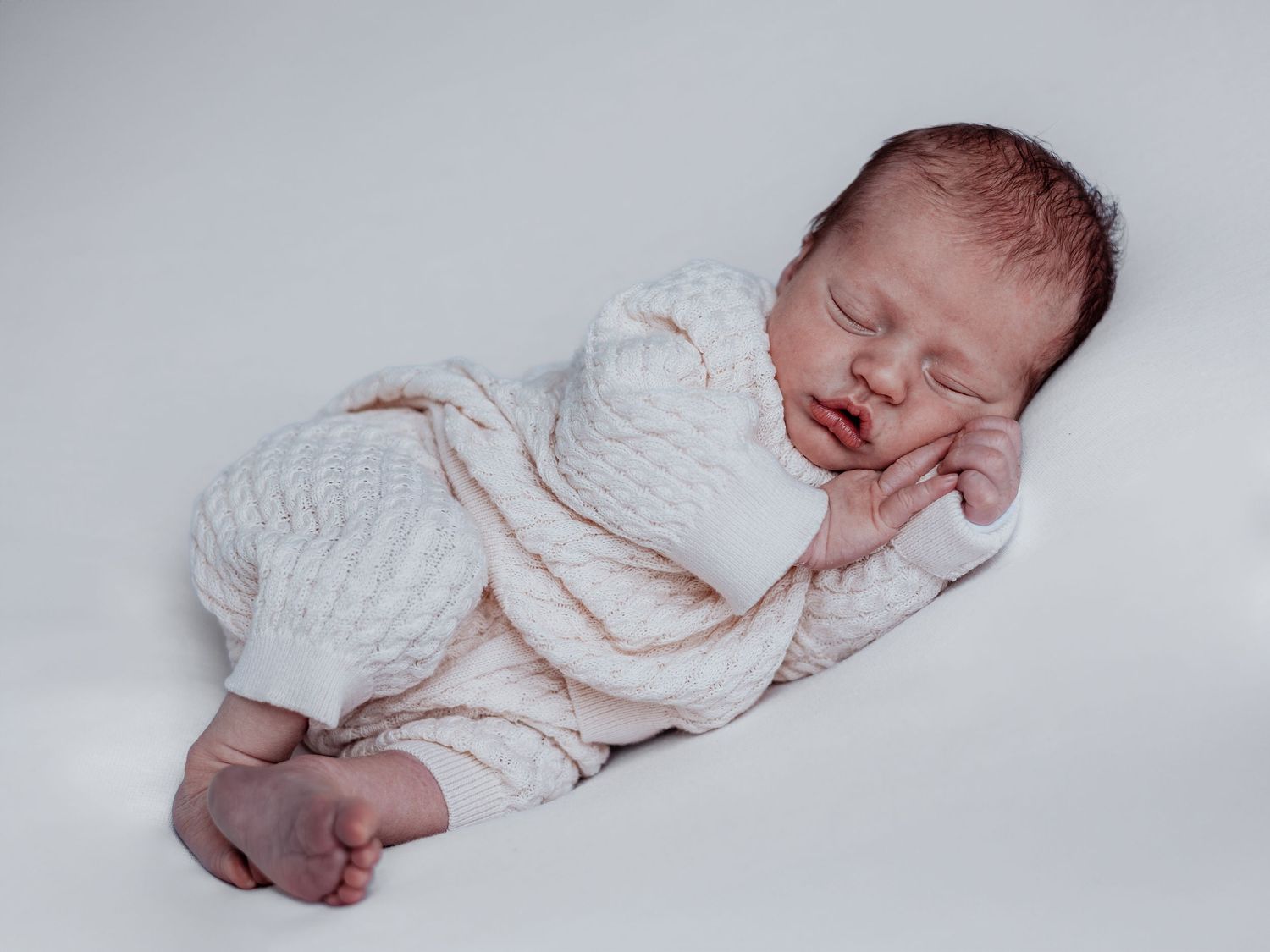 A sleeping newborn baby in white swaddling blanket rests peacefully on a light colored background.