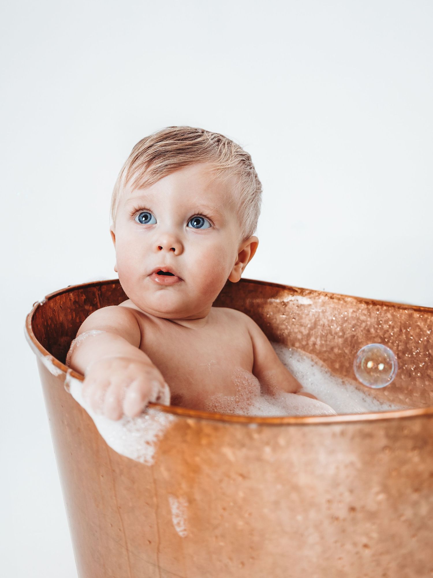 Blue-eyed baby sitting in a copper bathtub with bubbles, looking curiously at the camera.