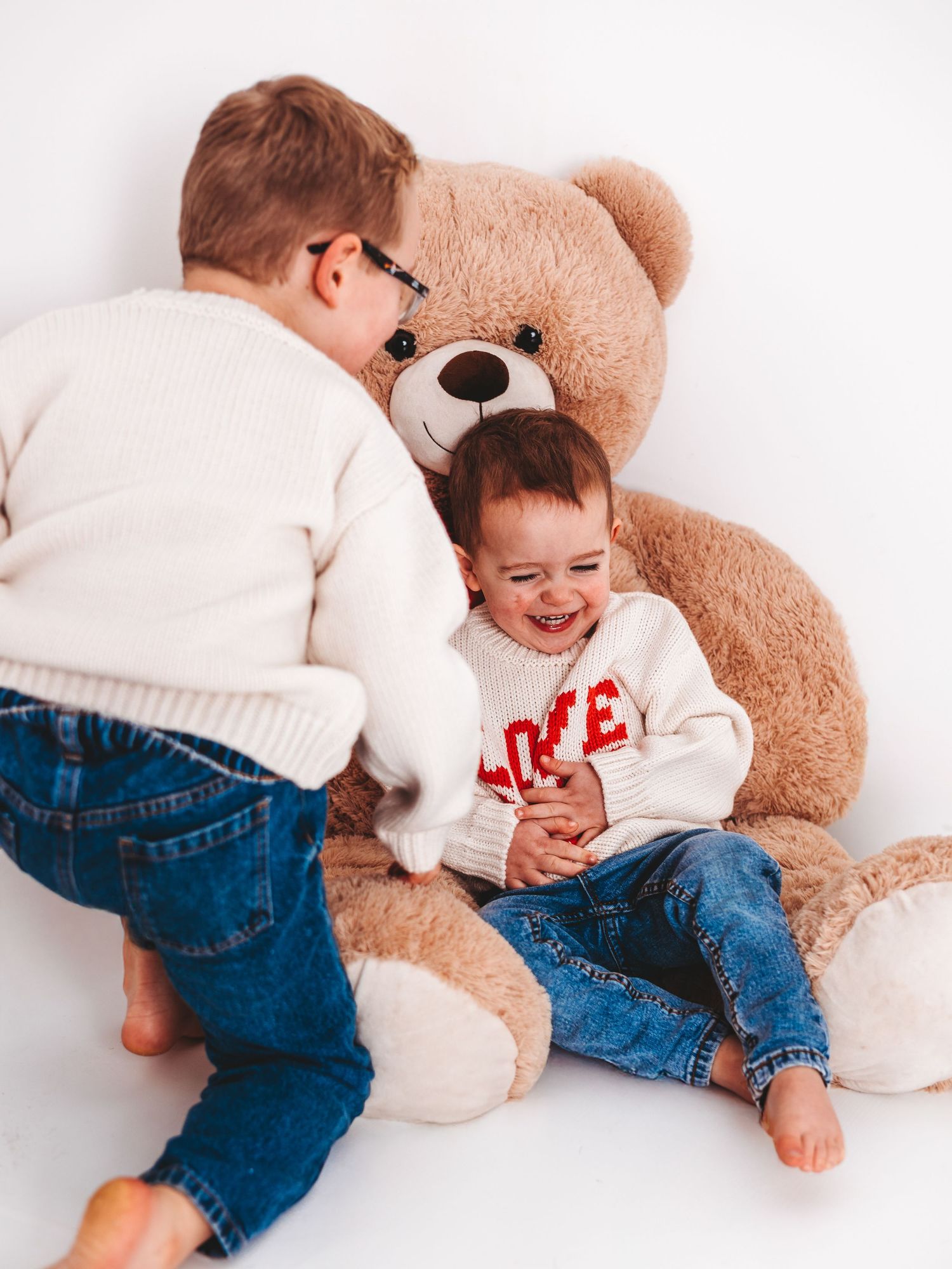 Two young children in white sweaters and jeans laugh together beside a large brown teddy bear on white background.