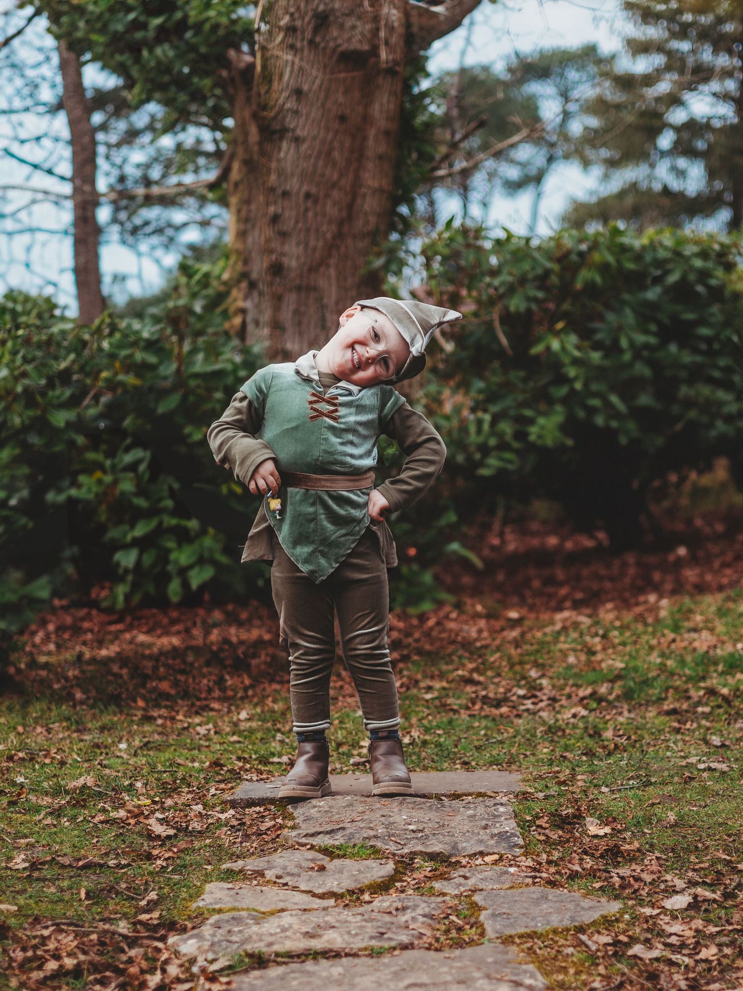 A young child dressed in a green elf costume poses confidently on a stone path in an autumn garden.
