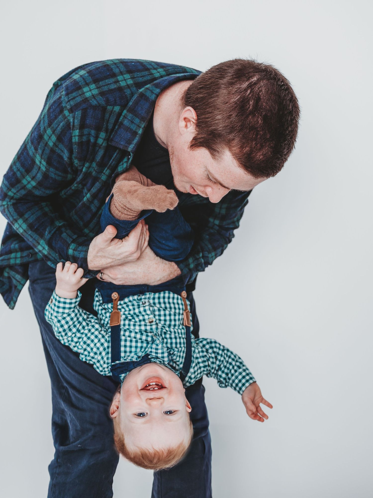 A playful father holds his laughing toddler upside down, both wearing plaid shirts, against a white background.