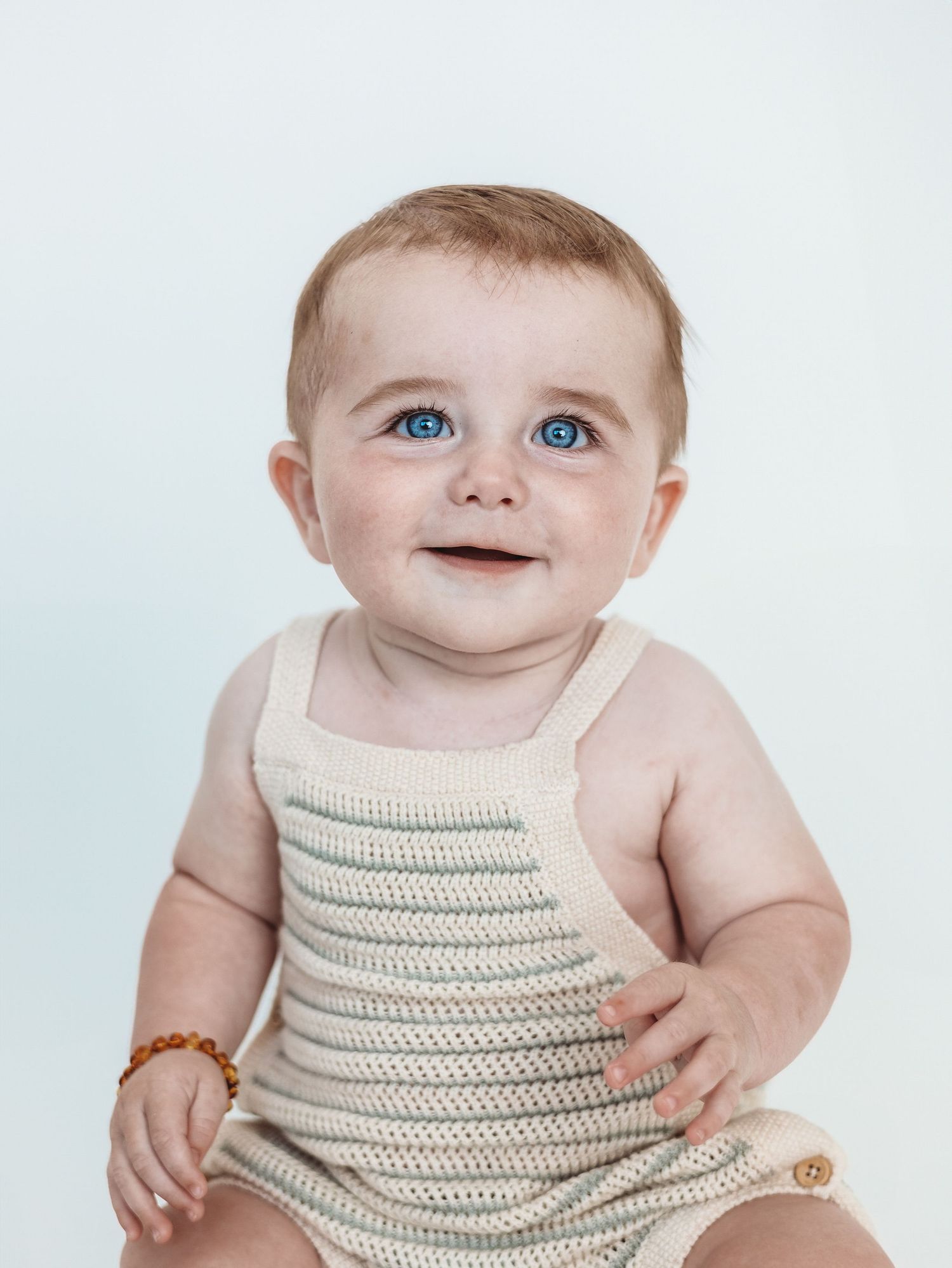 A baby wearing a cream-colored knit tank top smiles while sitting against a white background.