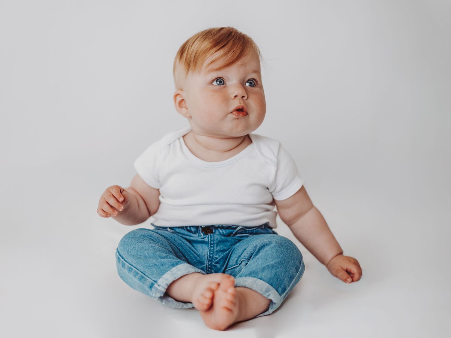 Baby wearing white t-shirt and blue jeans sits on white background looking up with curious expression.