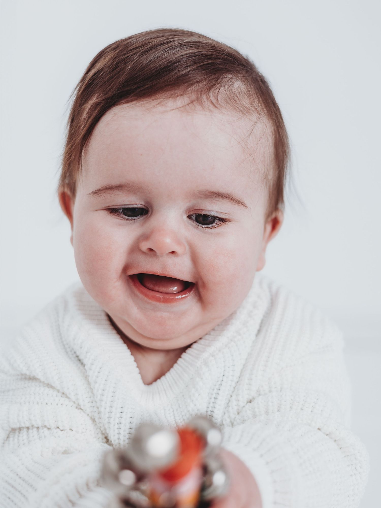 A happy baby wearing a white knit sweater smiles while reaching for something out of frame.