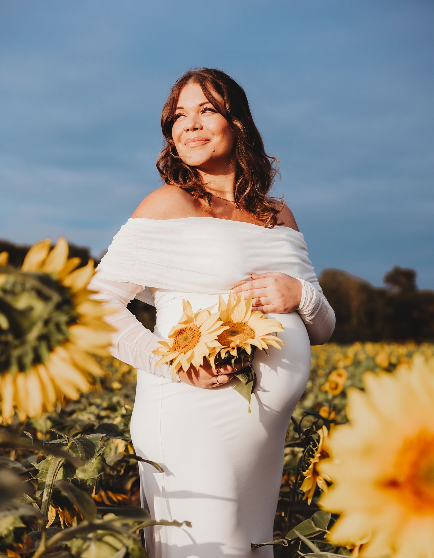 A person in a white dress stands in a golden sunflower field at sunset holding a bright orange sunflower bloom.