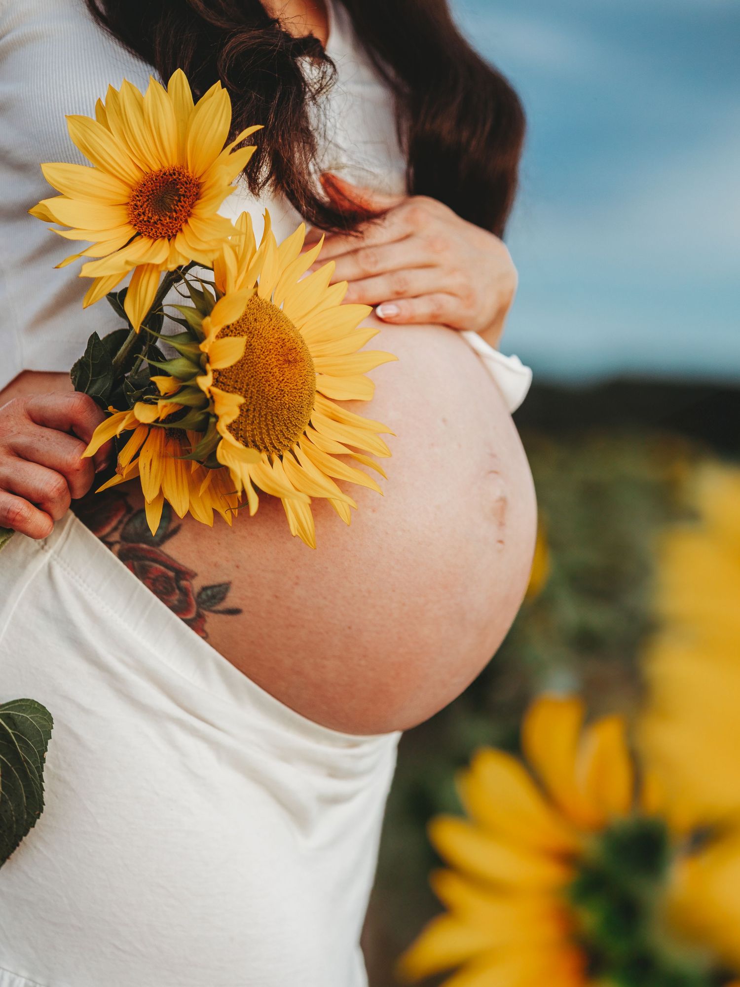 A pregnant woman in white dress stands in a sunflower field holding bright yellow blooms against a blue sky background.