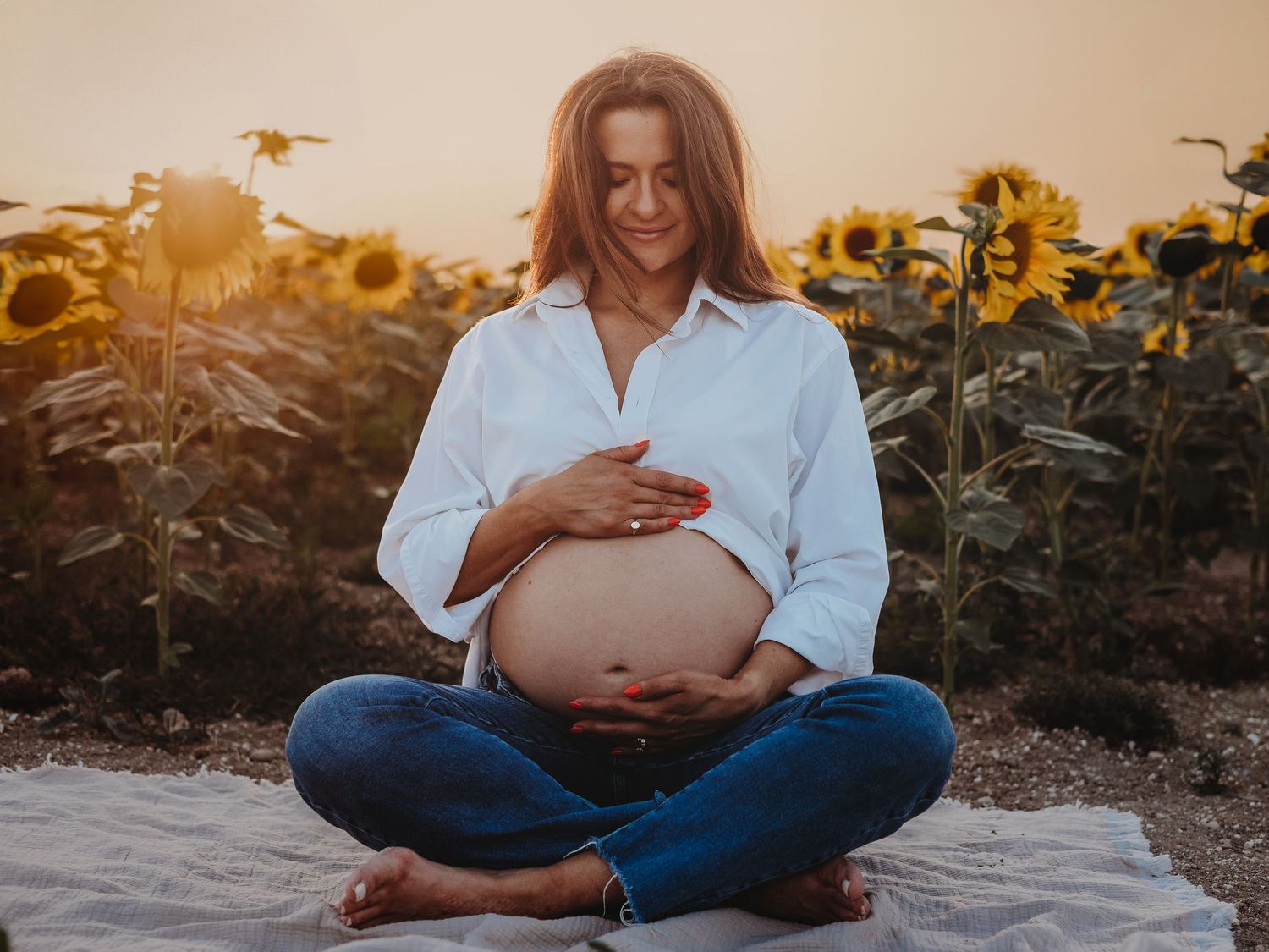 A maternity photo session at sunset in a sunflower field shows a person in white shirt and jeans touching their belly.