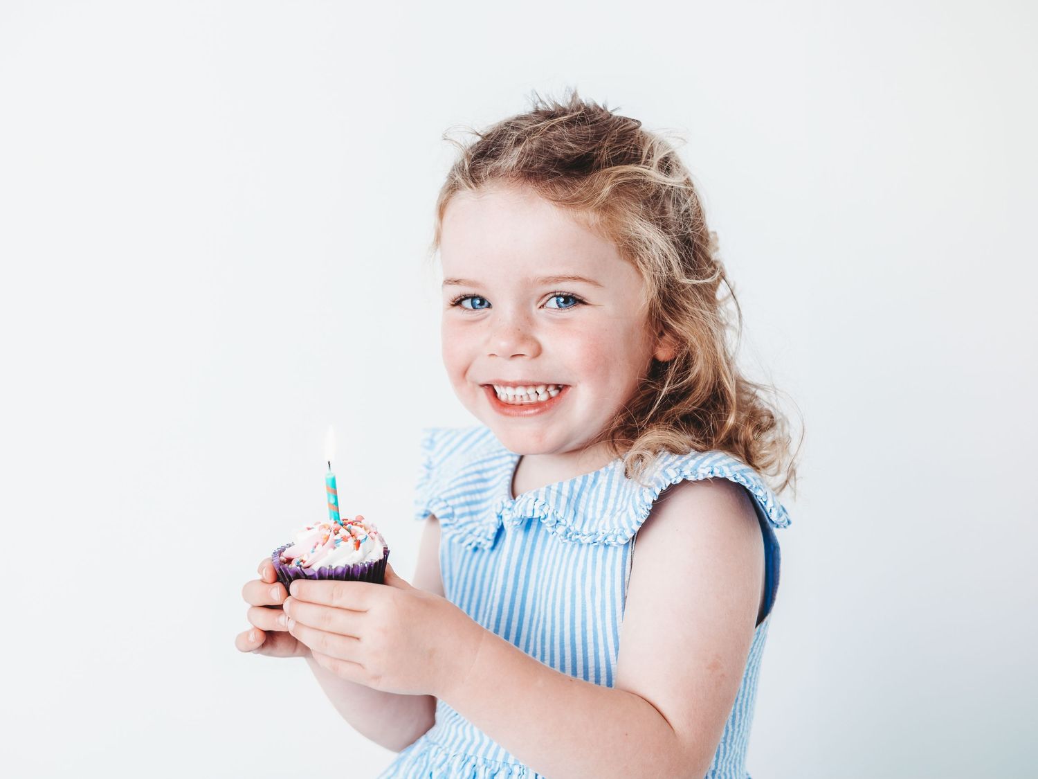 Happy young girl in blue dress holding a birthday cupcake with a candle, smiling against white background.