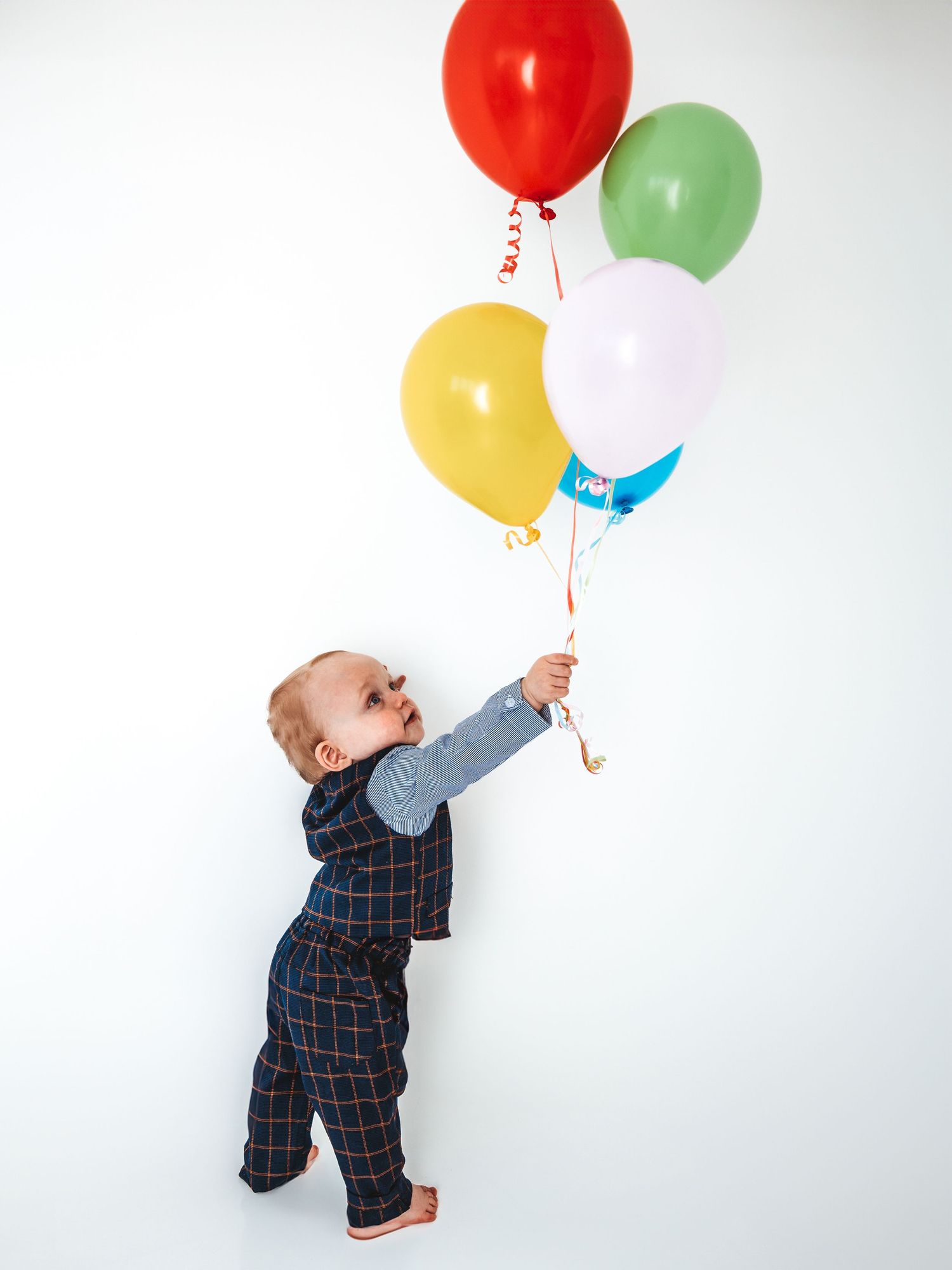 A curious baby in a plaid outfit holds a bunch of colorful balloons against a white background.