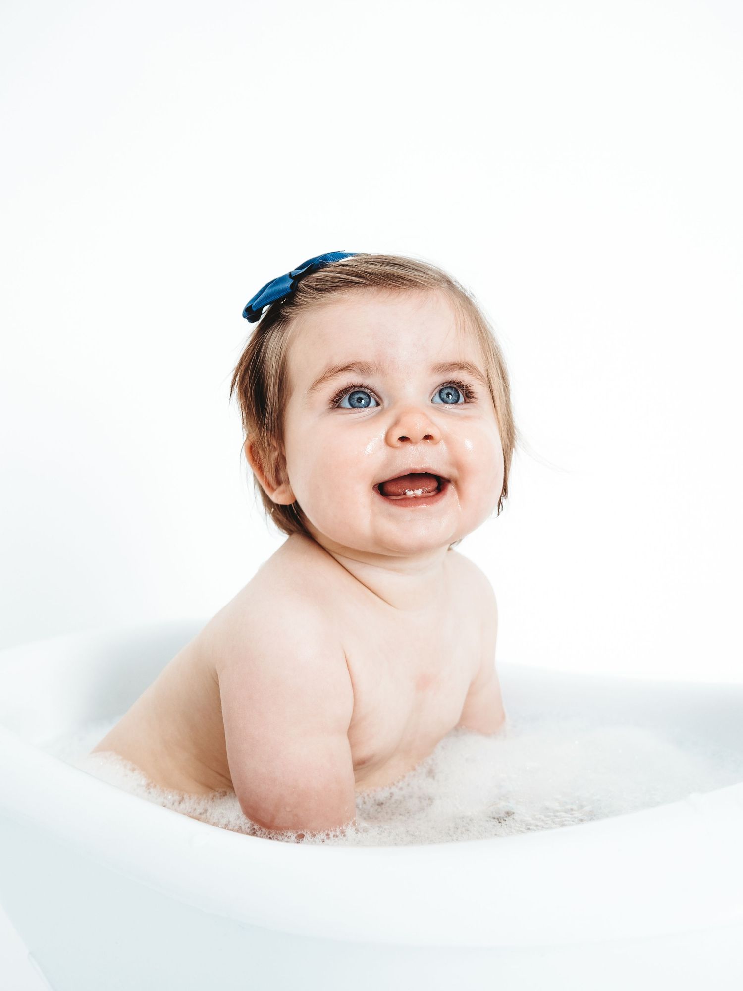 Smiling baby girl with blue bow sits in bubble bath, looking up with bright blue eyes against white background.