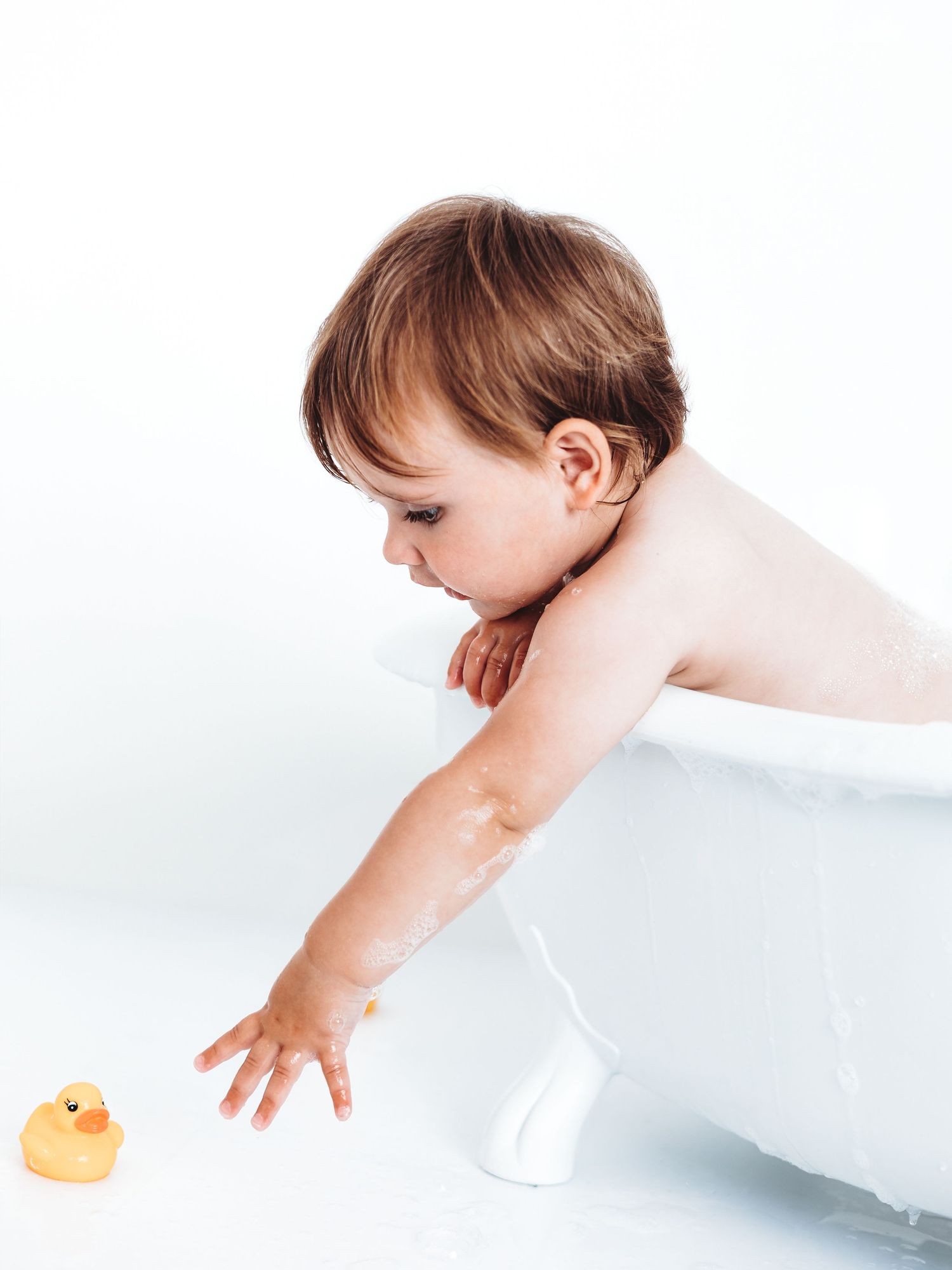 Baby in a white bathtub reaching for a yellow rubber duck, covered in soap suds during bath time.