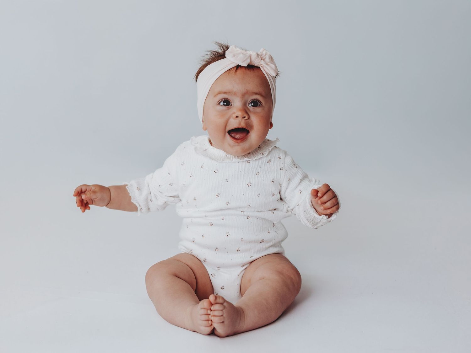 Smiling baby girl in white onesie and bow headband sitting on light gray background.