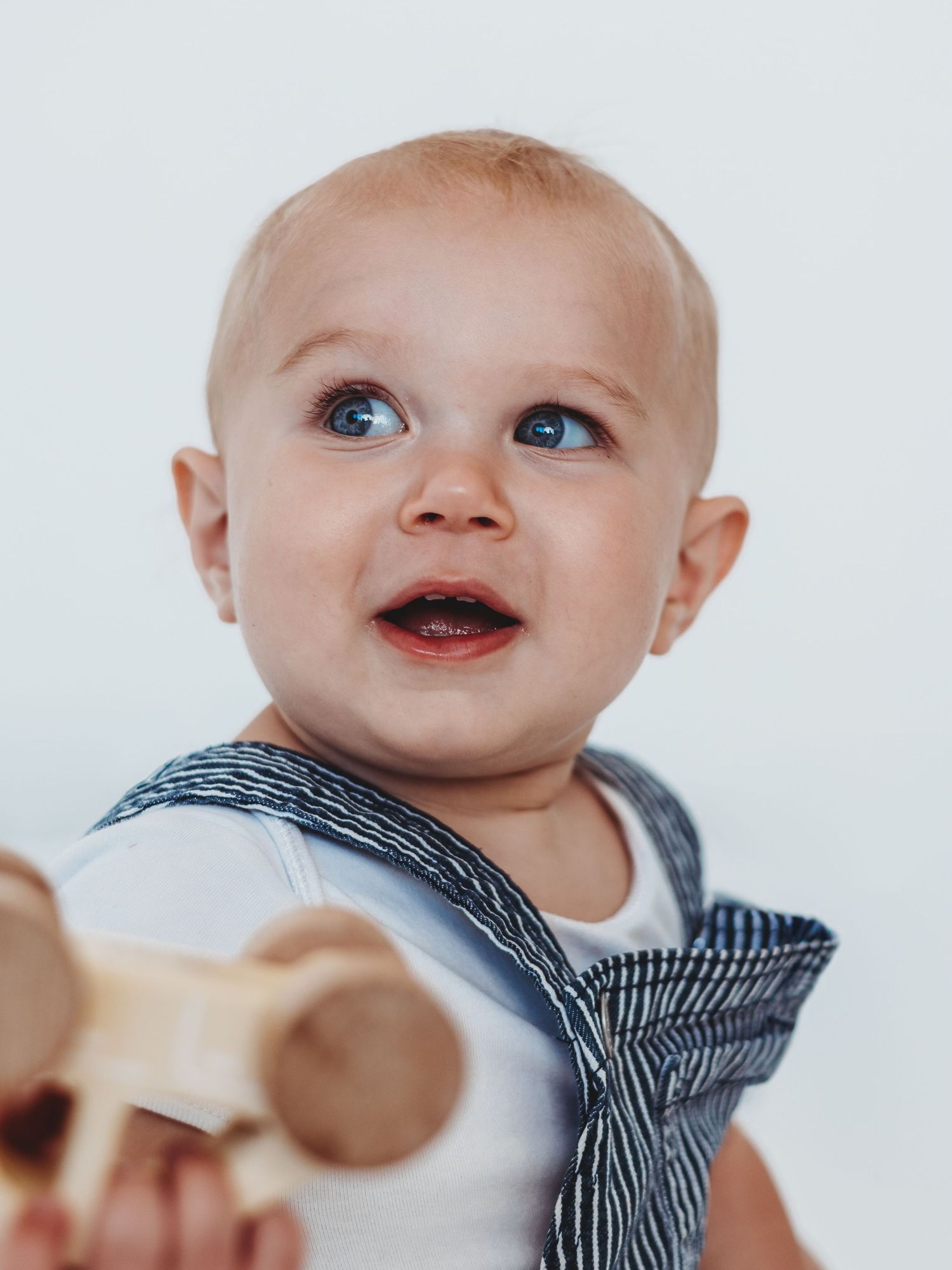 A baby in striped clothing smiles while holding a wooden spoon during mealtime.