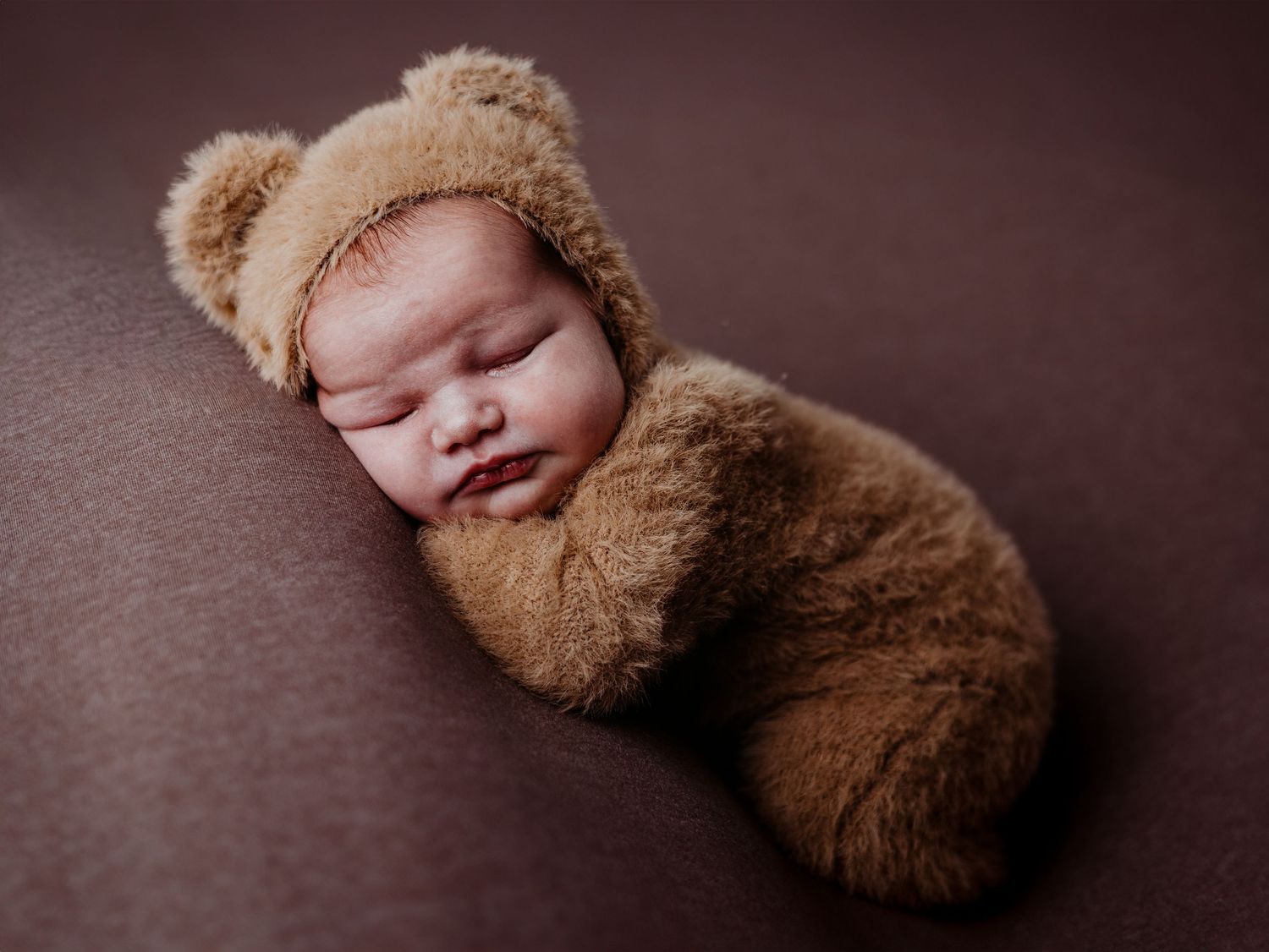 A sleeping baby wrapped in a brown teddy bear outfit lies on a brown textured surface.