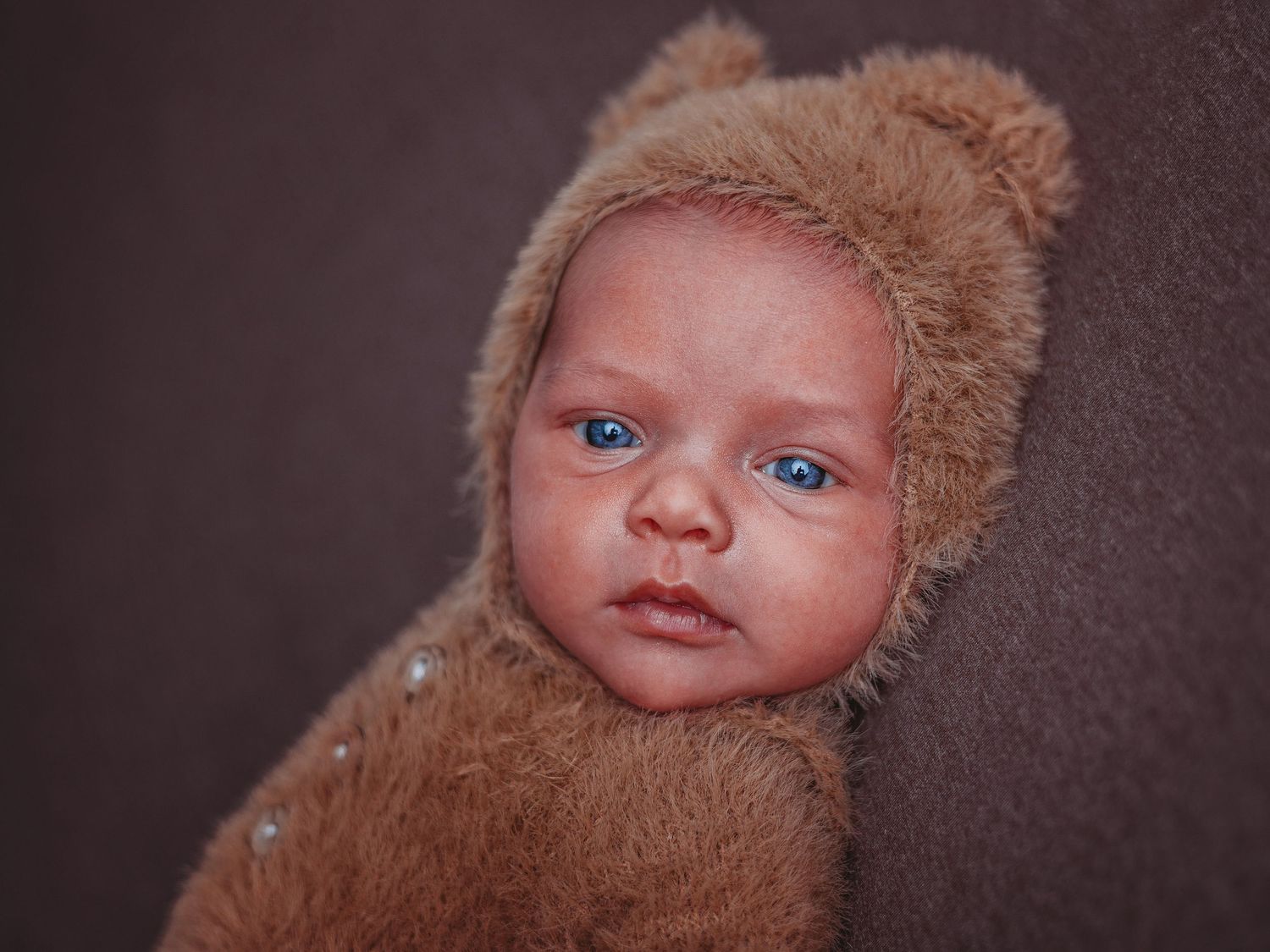 A baby wearing a soft brown teddy bear style outfit with bright blue eyes rests against a dark background.