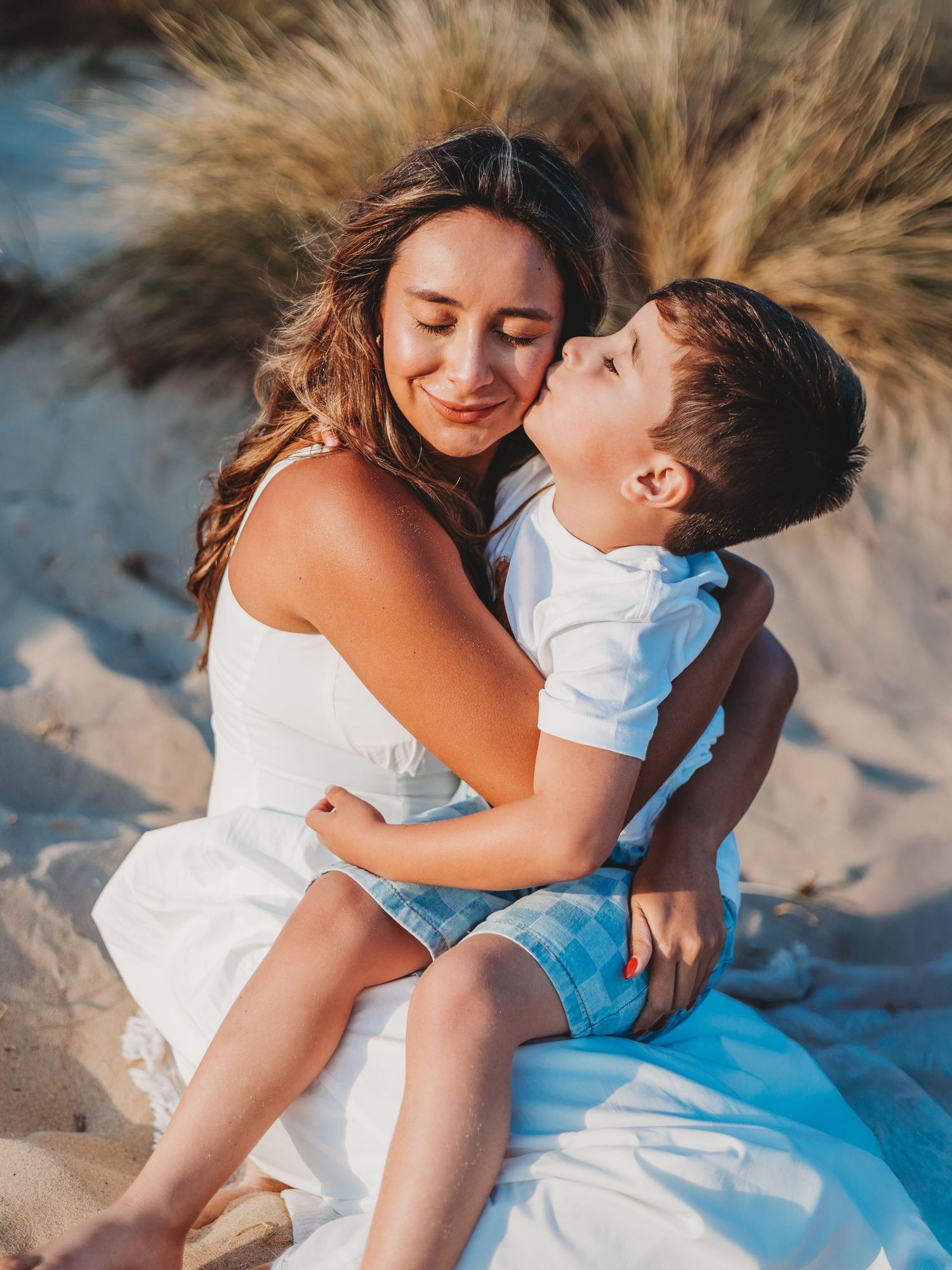 A woman in a white dress sits on sandy beach dunes while a young boy kisses her cheek affectionately.
