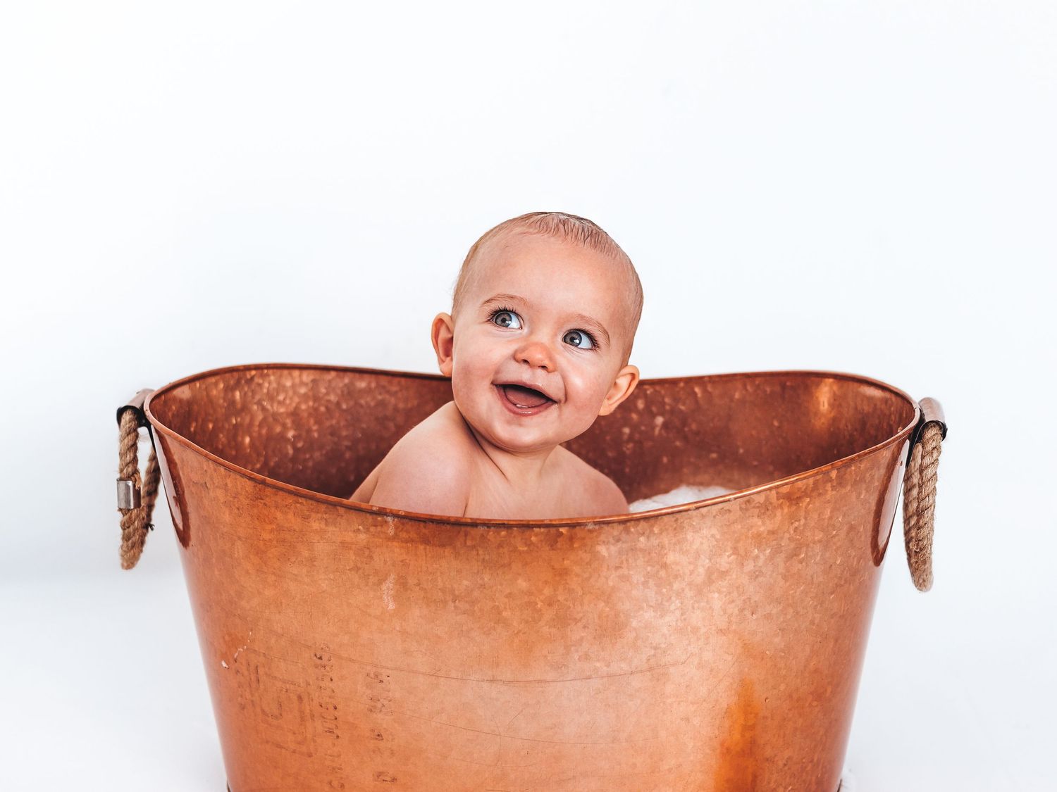 Smiling baby sitting in a rustic copper bathtub against a white background.