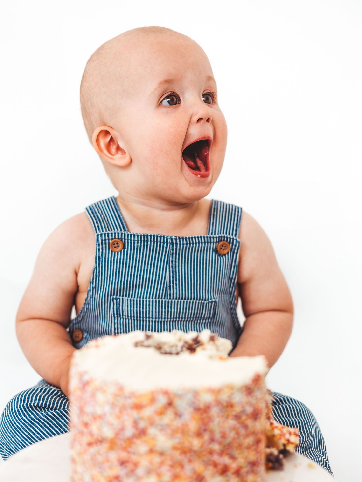 Excited baby in denim overalls with mouth open wide in front of a colorful smash cake on white background.