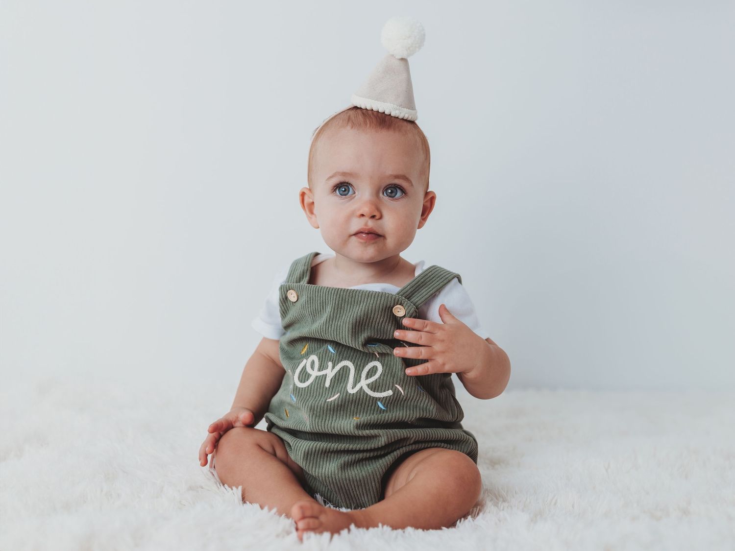 Baby wearing olive green romper with 'one' text sits on white fluffy rug looking upward.