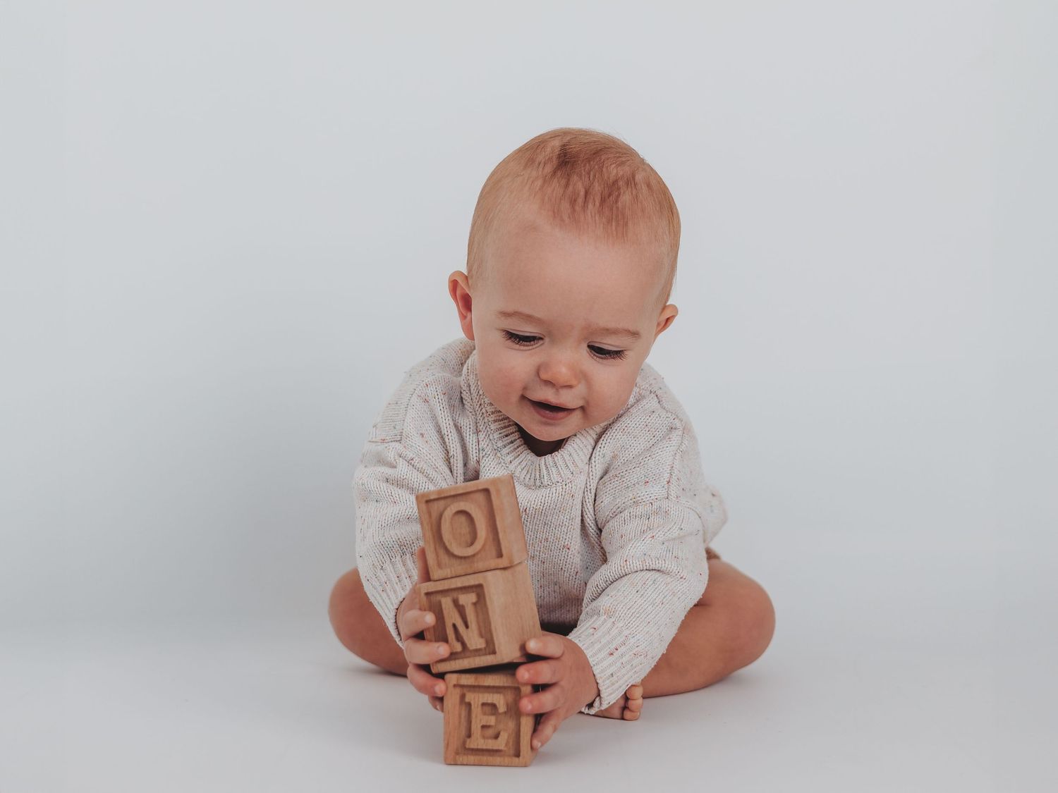 A baby in white clothing lies on a light background playing with wooden alphabet blocks.