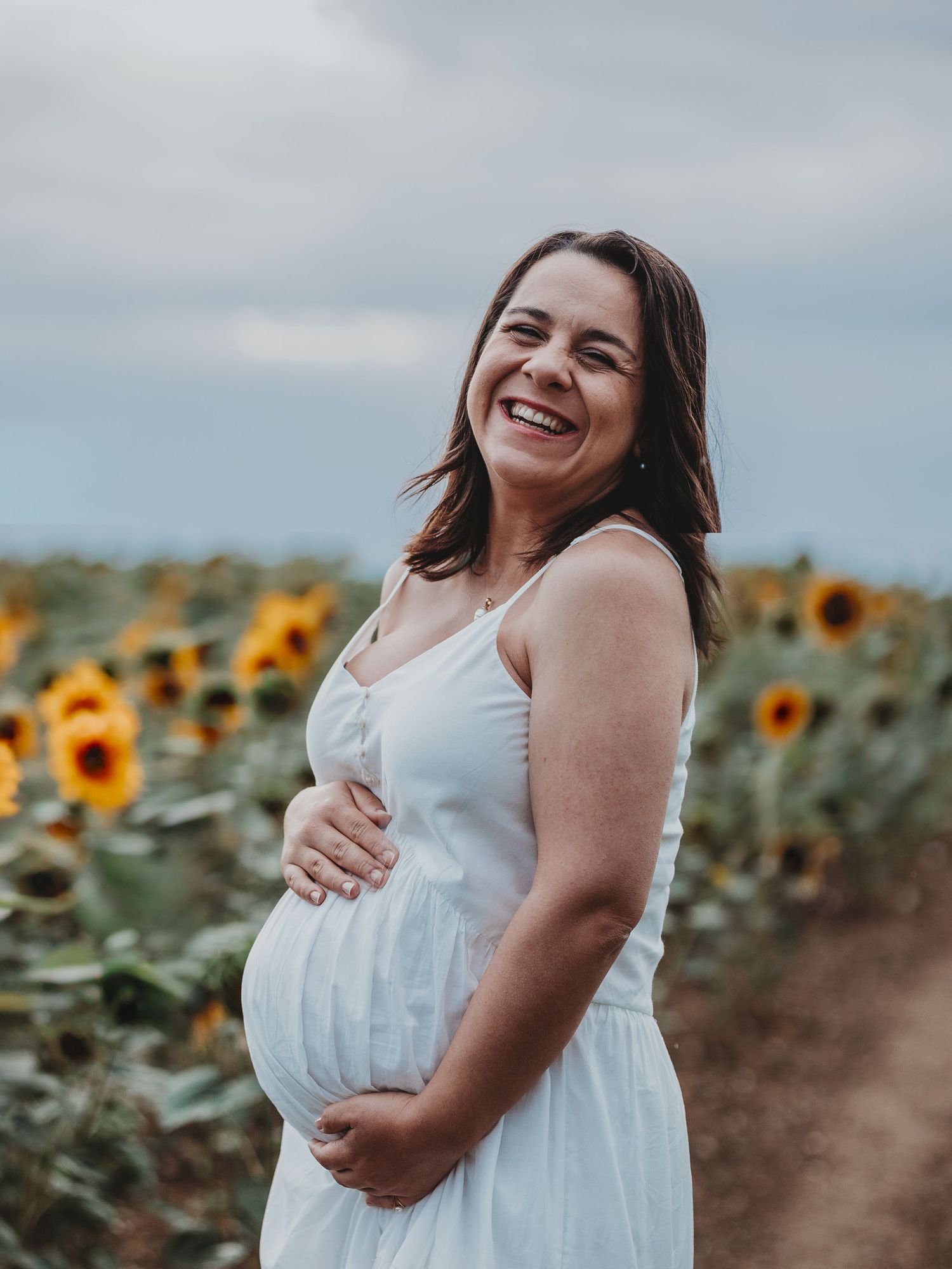 A person in a white dress stands joyfully among blooming sunflowers during a maternity photoshoot at sunset.