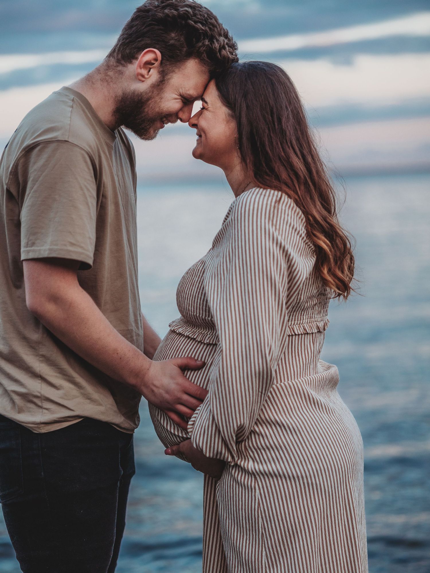 Expectant couple shares an intimate moment by the water during sunset, with the pregnant woman wearing a striped dress.