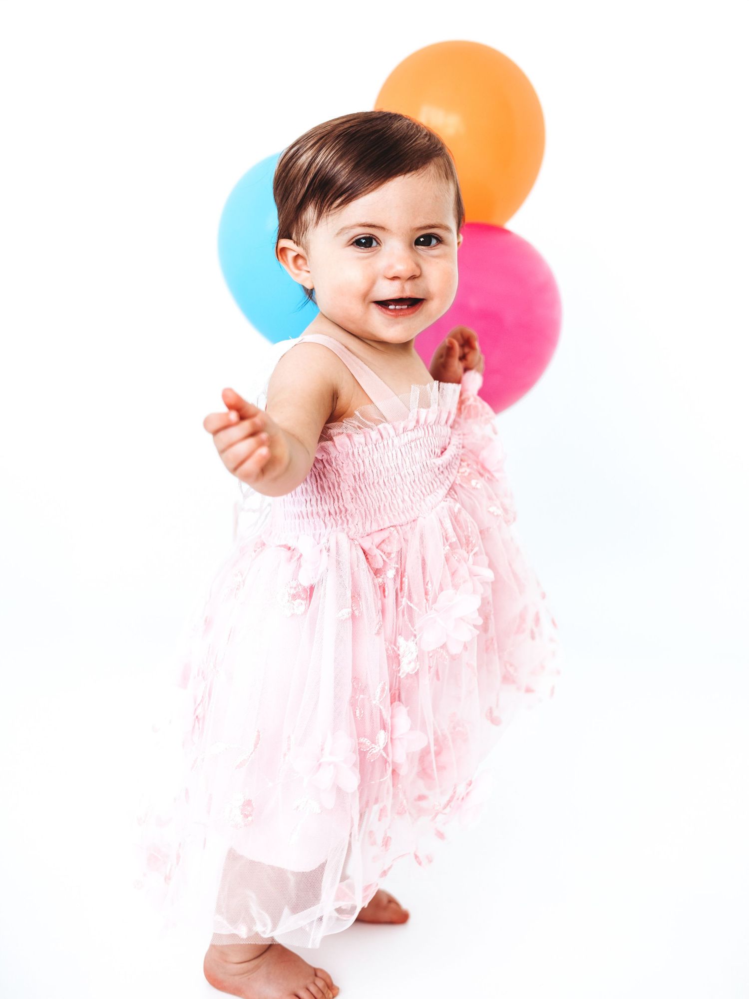 A smiling baby girl in a pink dress stands barefoot holding colorful balloons on a white background.